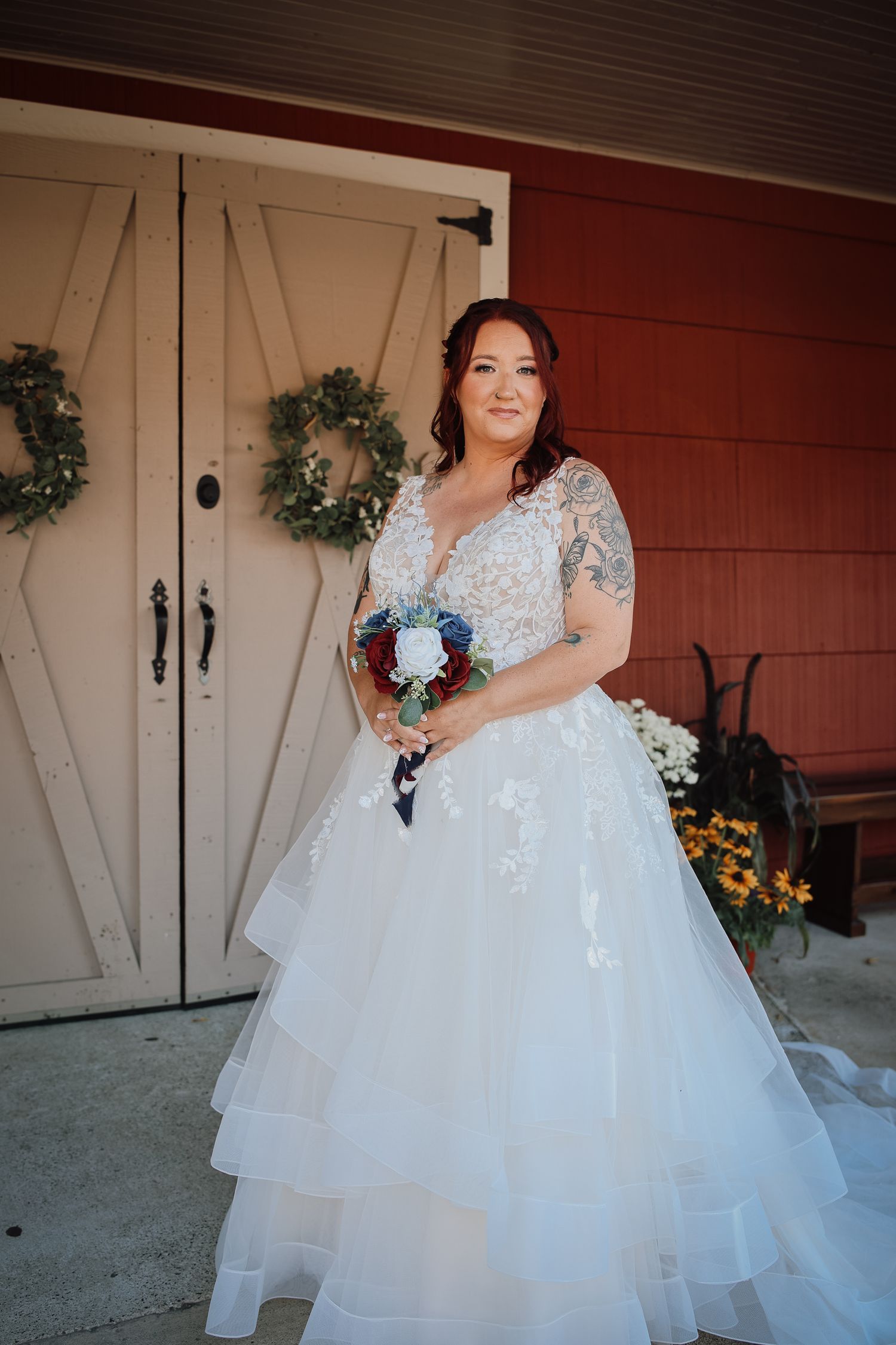 A bride in a white lace wedding gown with layered tulle skirt holds a bouquet in front of rustic barn doors with wreaths.