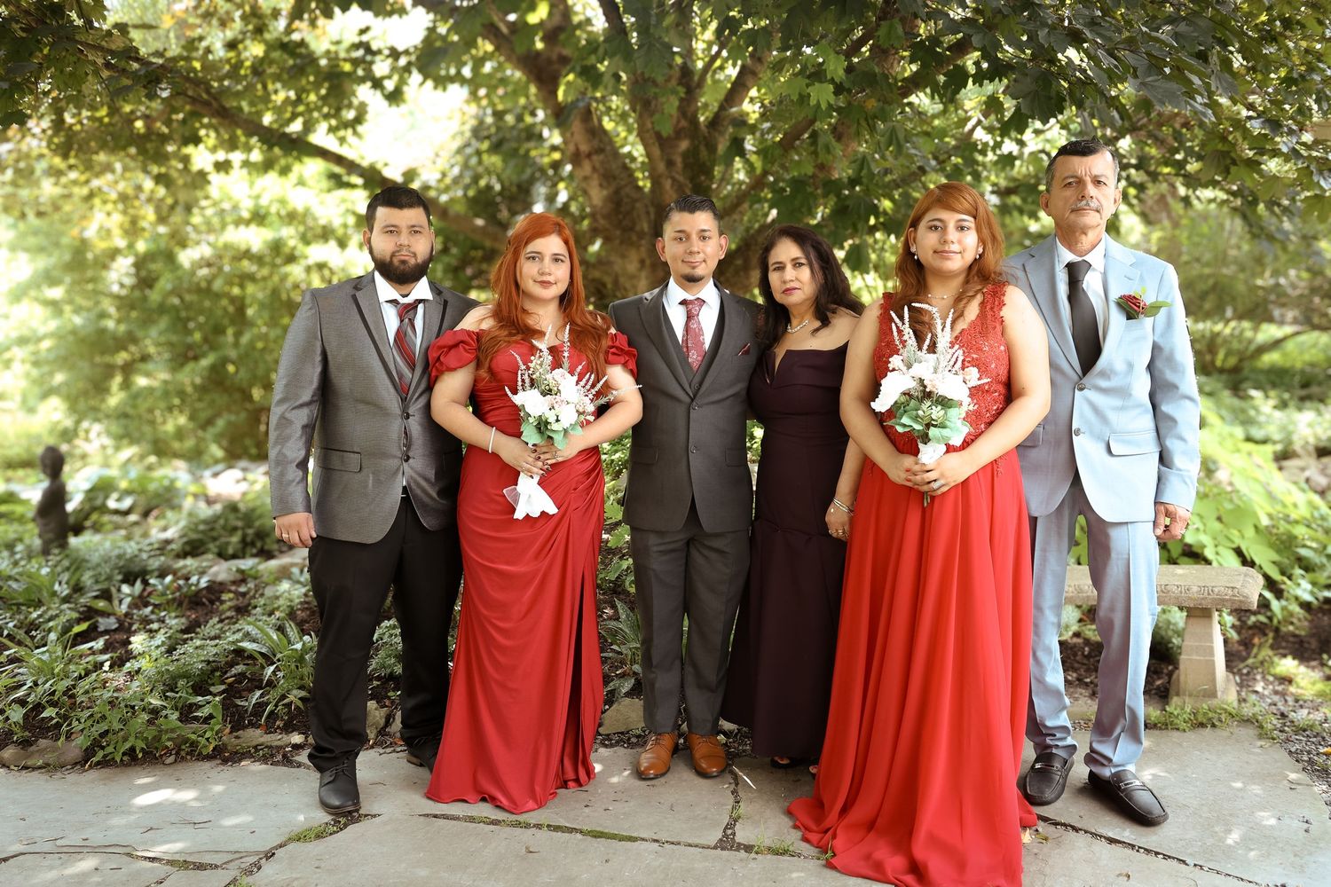Wedding party in red dresses and dark suits poses together outdoors in a garden setting.