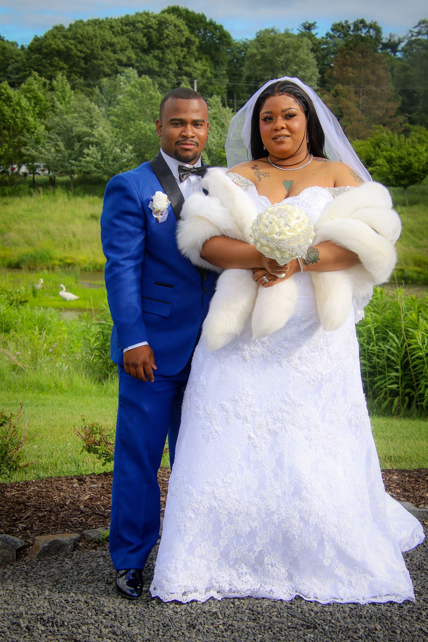 A newlywed couple poses outdoors in formal wedding attire with a blue suit and white lace gown against a green landscape.