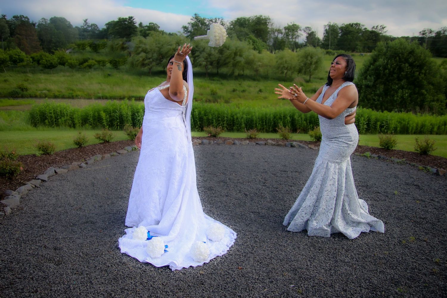 Two people in white dresses sit on a gravel path surrounded by green fields and trees on a cloudy day.