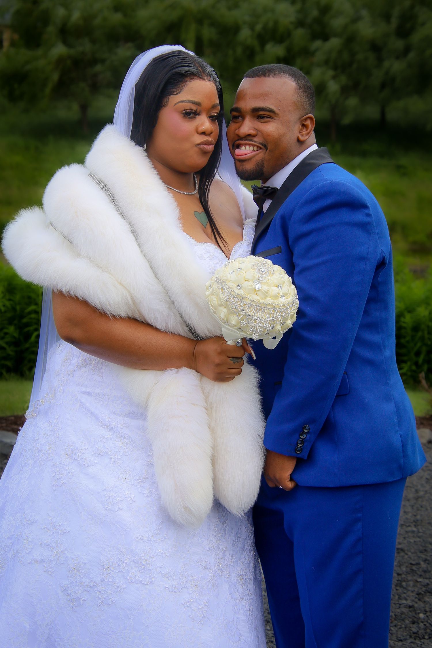 A bride in a white wedding dress with fur wrap holds a white bouquet while posing with groom in royal blue suit.
