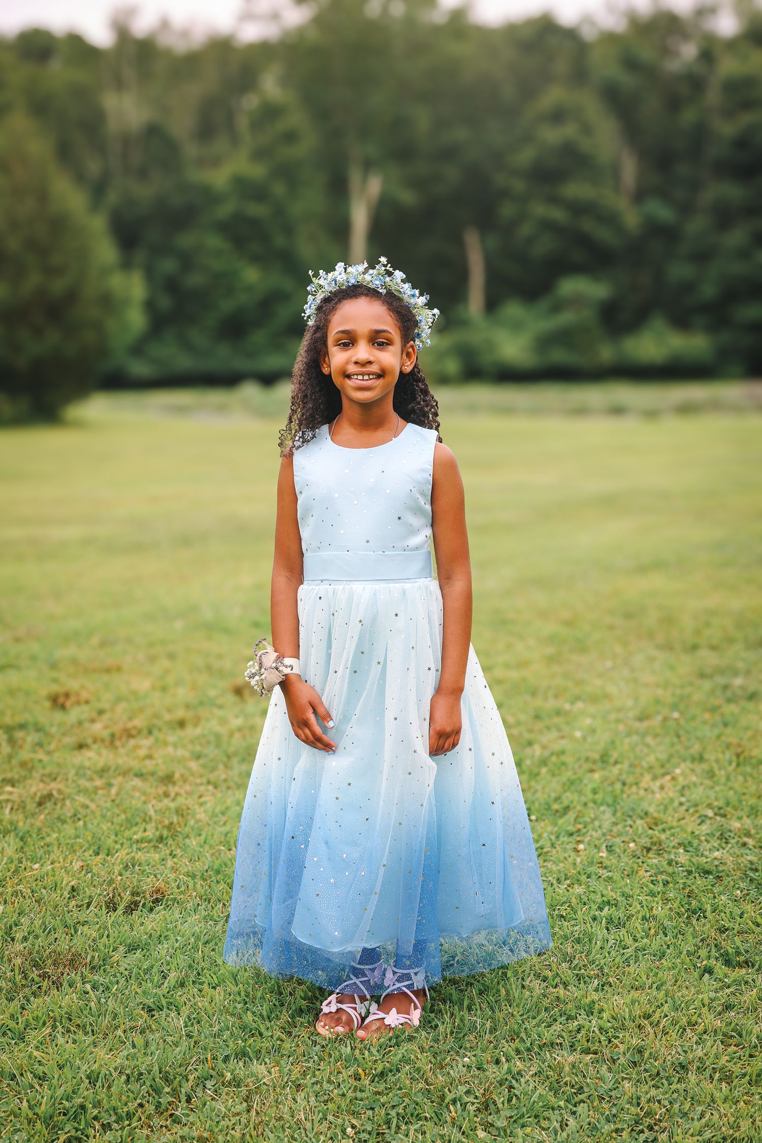 A child in a light blue tulle dress poses joyfully on a grassy lawn during a sunny day.