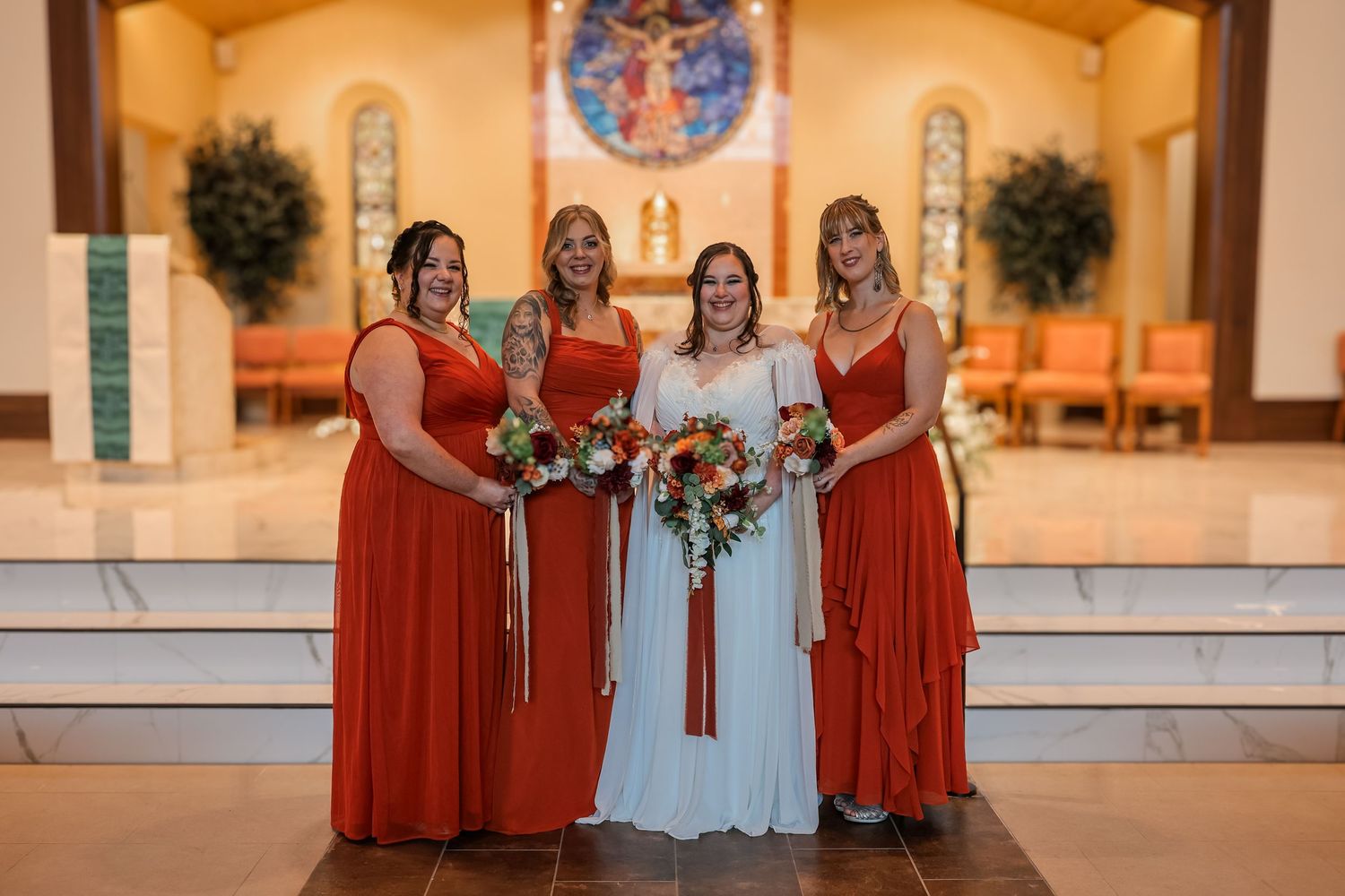 Wedding party members in burnt orange bridesmaid dresses pose with the bride in a white gown at a church ceremony.