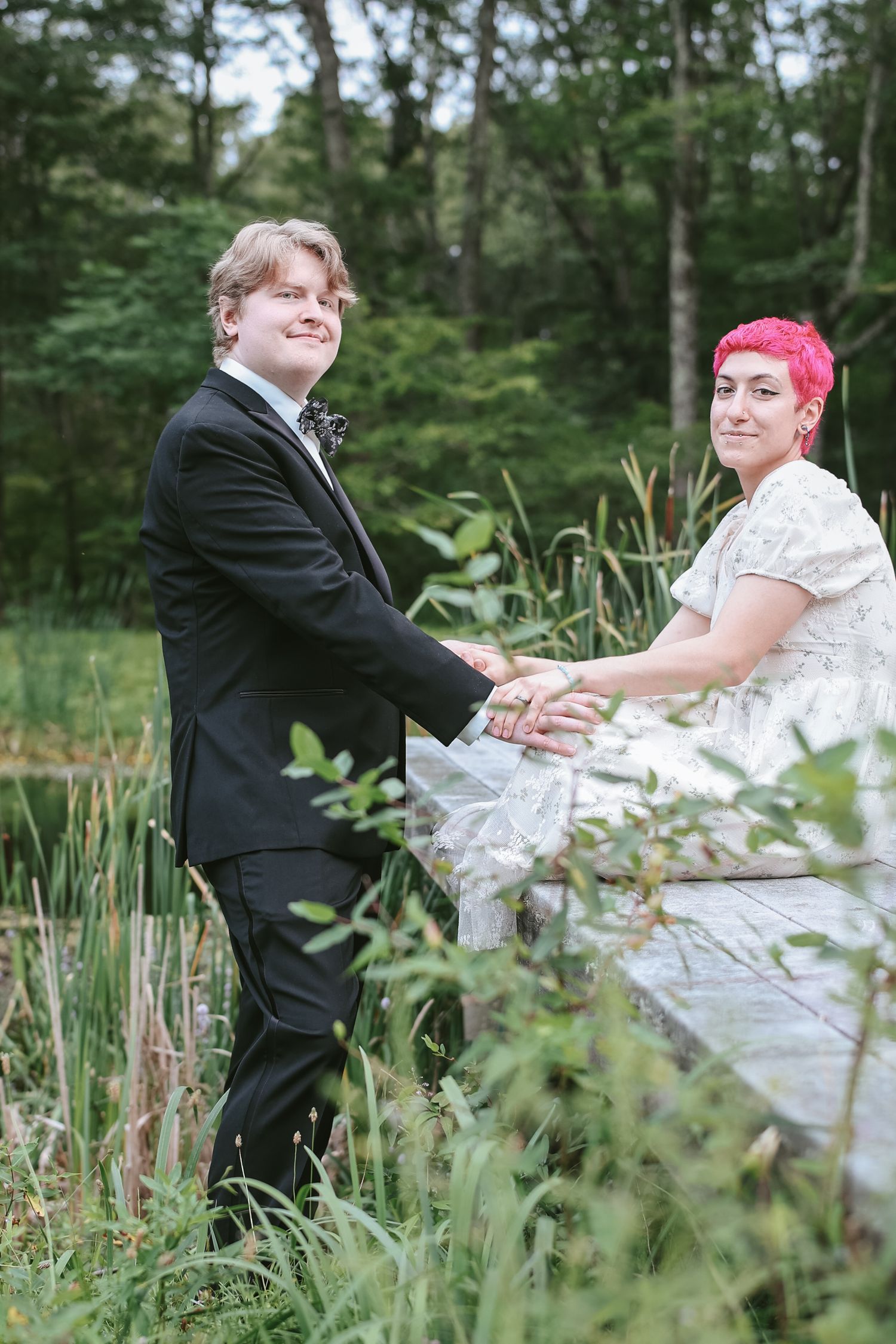 A couple in wedding attire pose together on a wooden boardwalk surrounded by lush greenery and cattails.