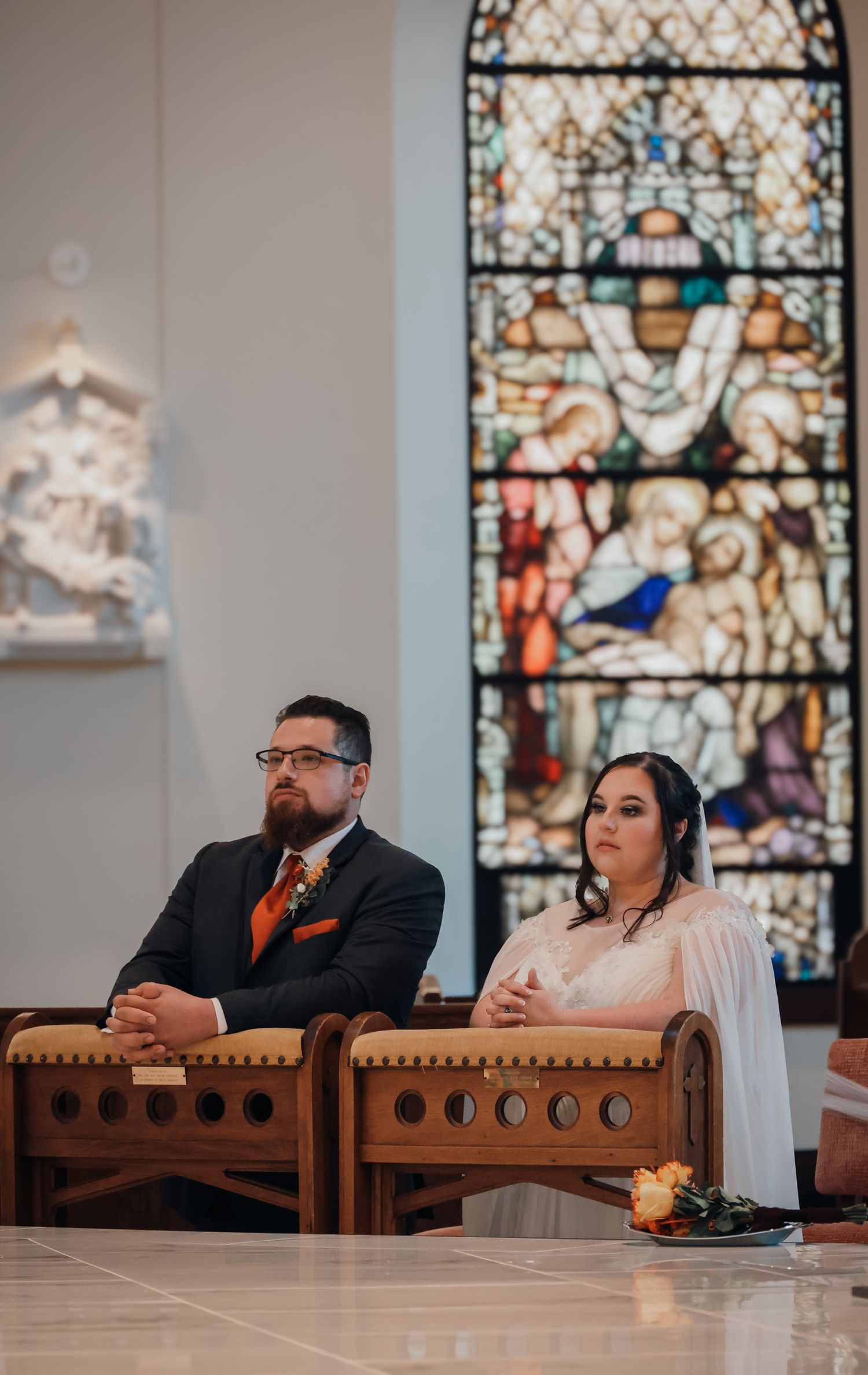 A couple sits in wooden church pews during a ceremony with stained glass windows in the background.