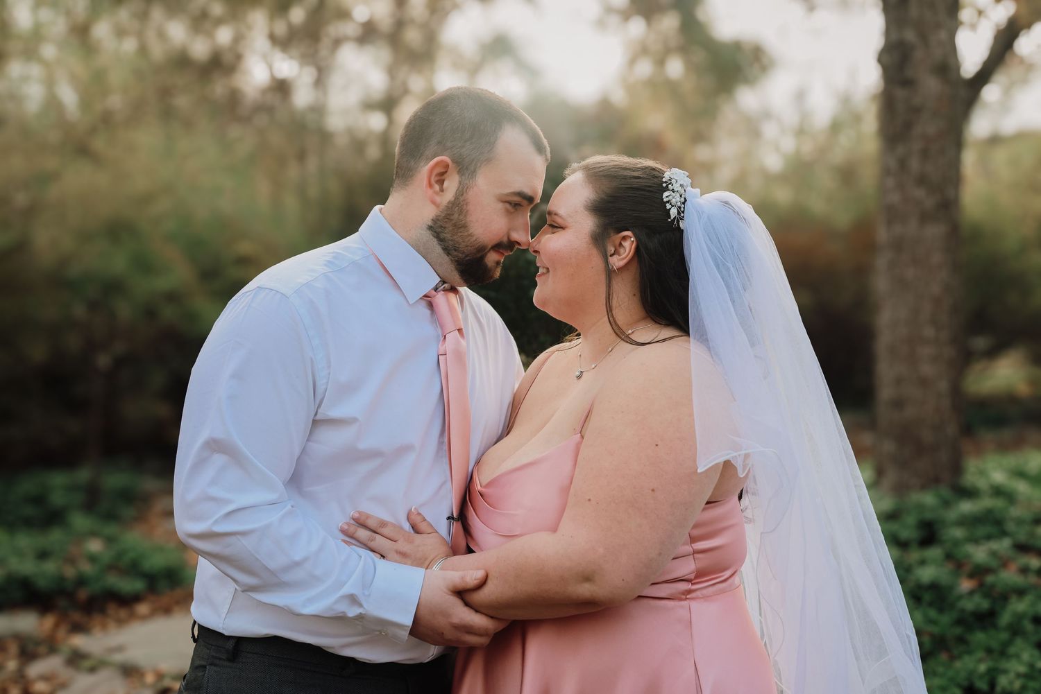 A couple in pink and white attire share an intimate moment outdoors during their wedding photoshoot.