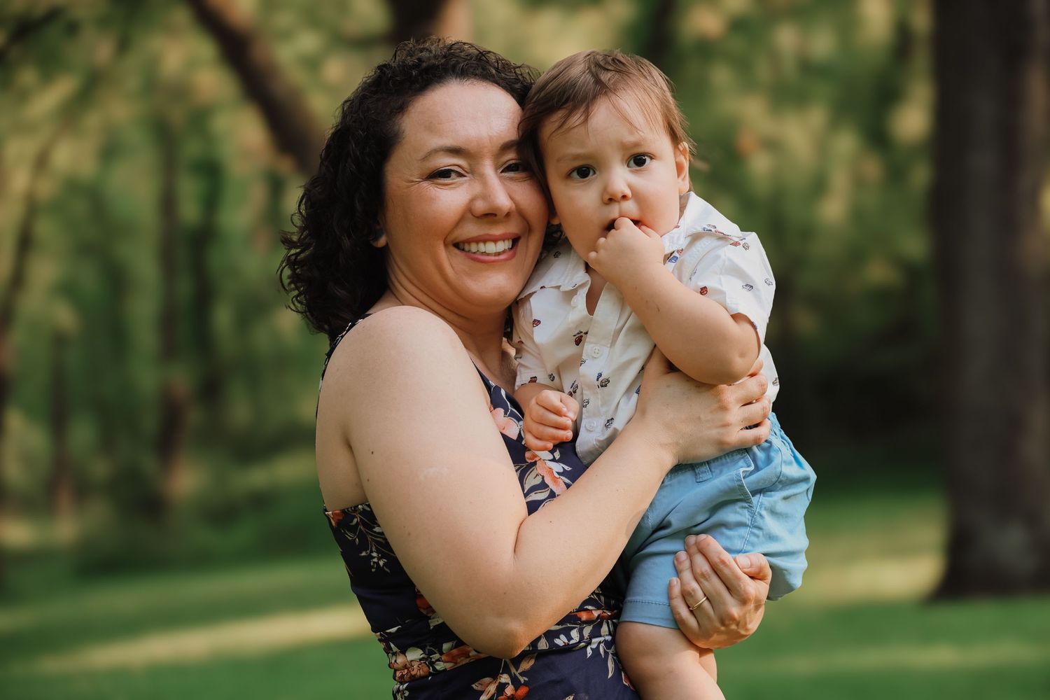 A floral dress-clad adult and toddler share a joyful moment together in a sunlit wooded setting.