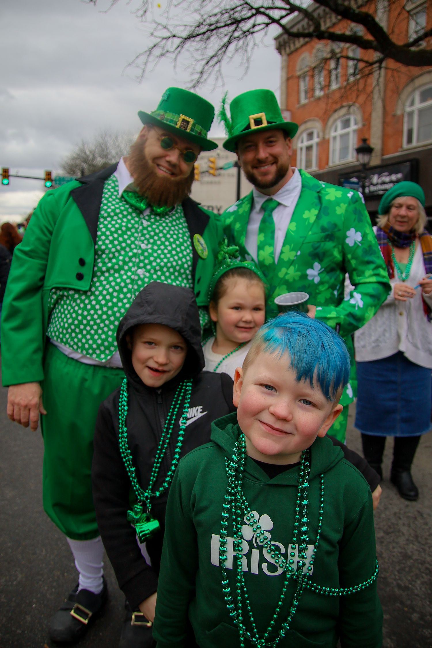 Festive parade goers wearing green outfits and beaded necklaces celebrate St. Patrick's Day on a cloudy afternoon.