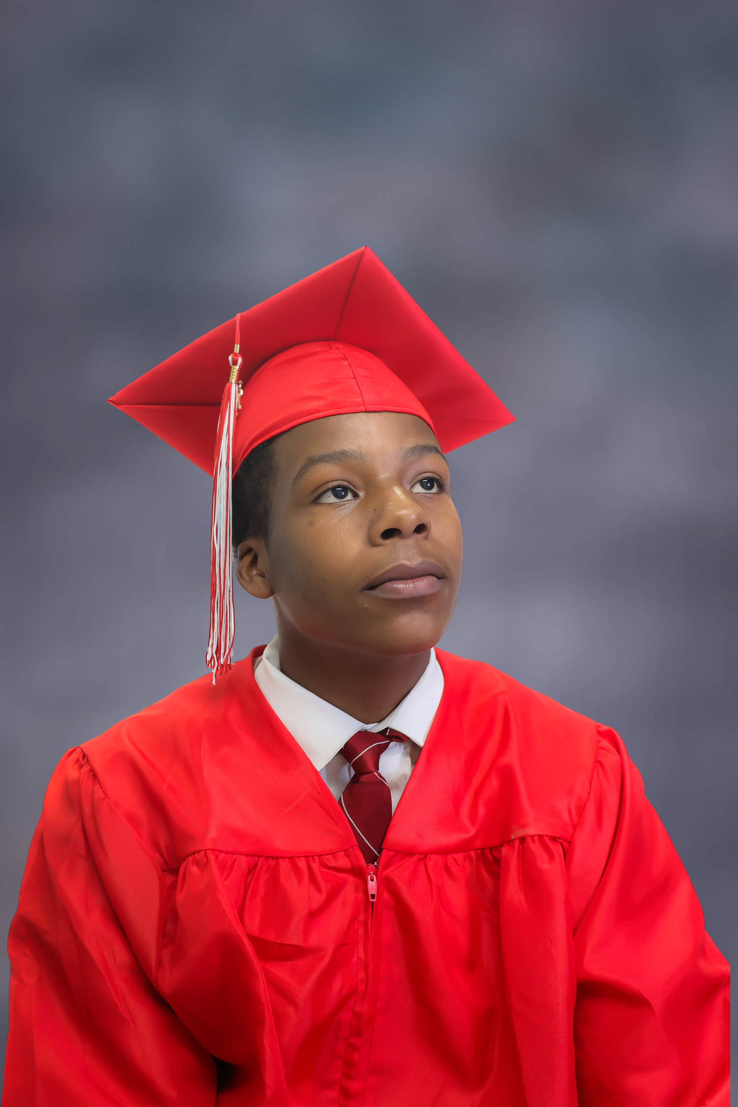 Graduate wearing bright red cap and gown with white tassel and red tie poses for portrait against gray background.