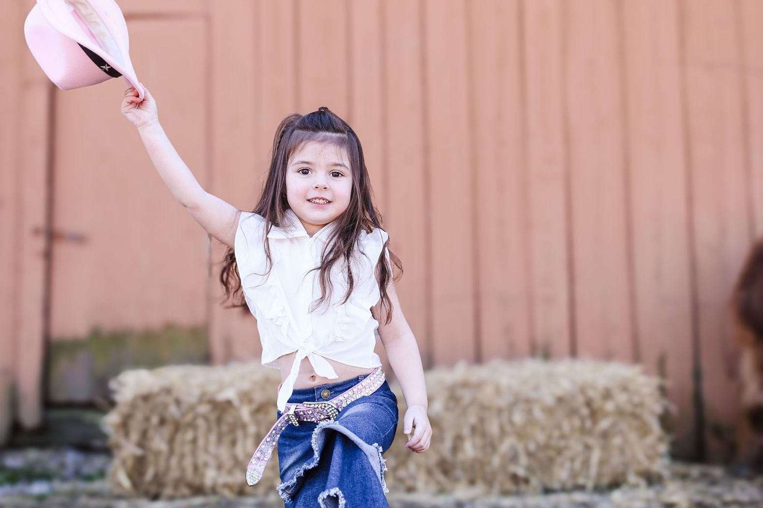 A young child in a blue dress twirls and dances in front of a rustic barn wall with hay bales visible.