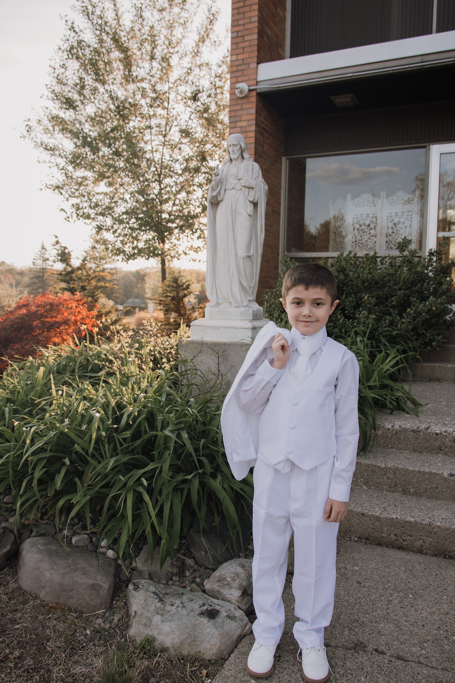 A figure in a white suit stands next to a religious statue and autumn foliage outside a brick building.
