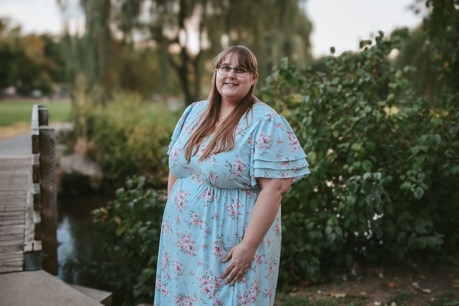 Woman in light blue floral dress poses outdoors in garden setting during golden hour.
