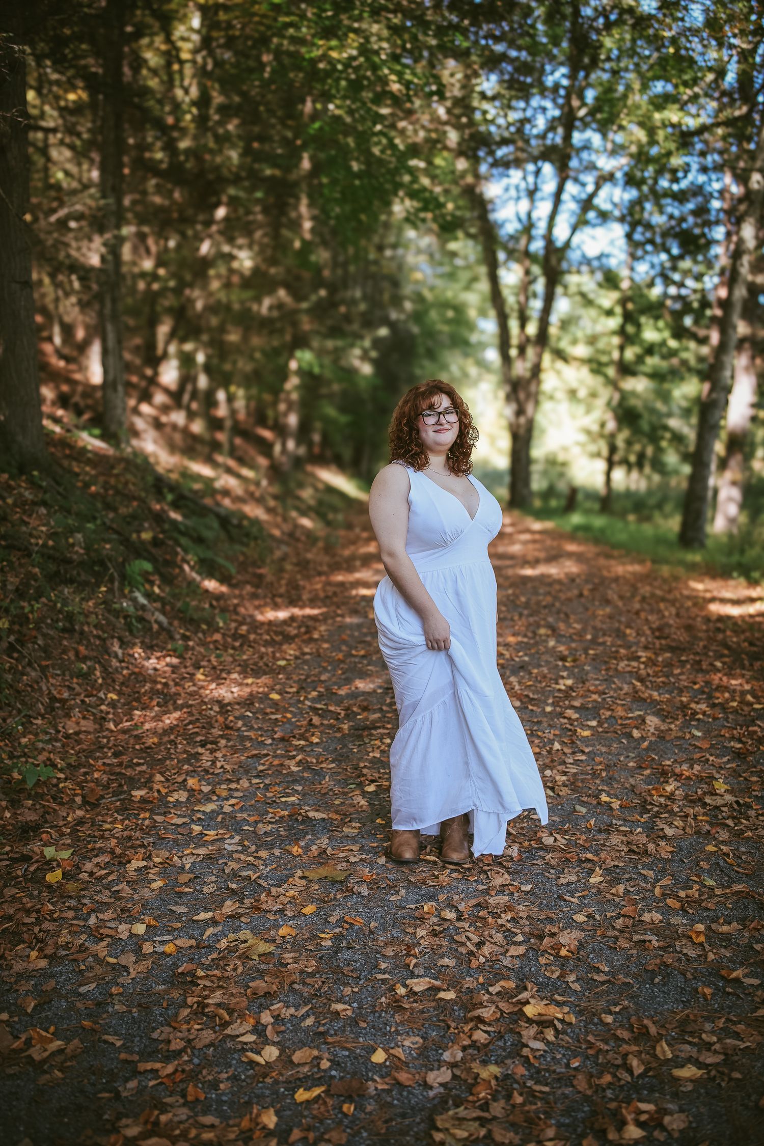 A person in a flowing white dress twirls gracefully among fallen autumn leaves on a wooded forest path.