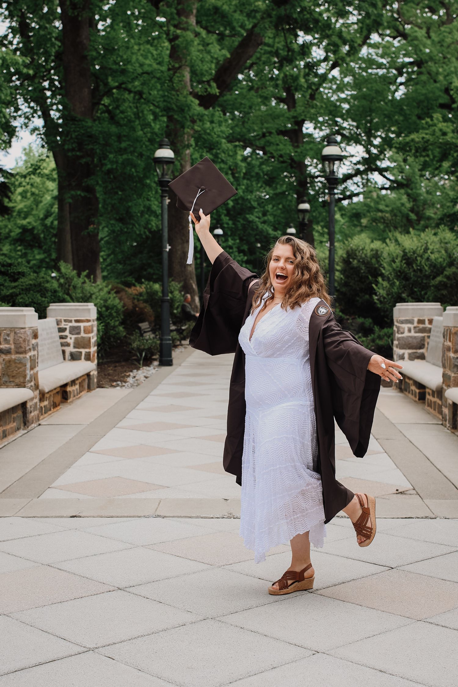 A graduate in a white dress and black graduation gown celebrates joyfully on a stone pathway surrounded by green trees.