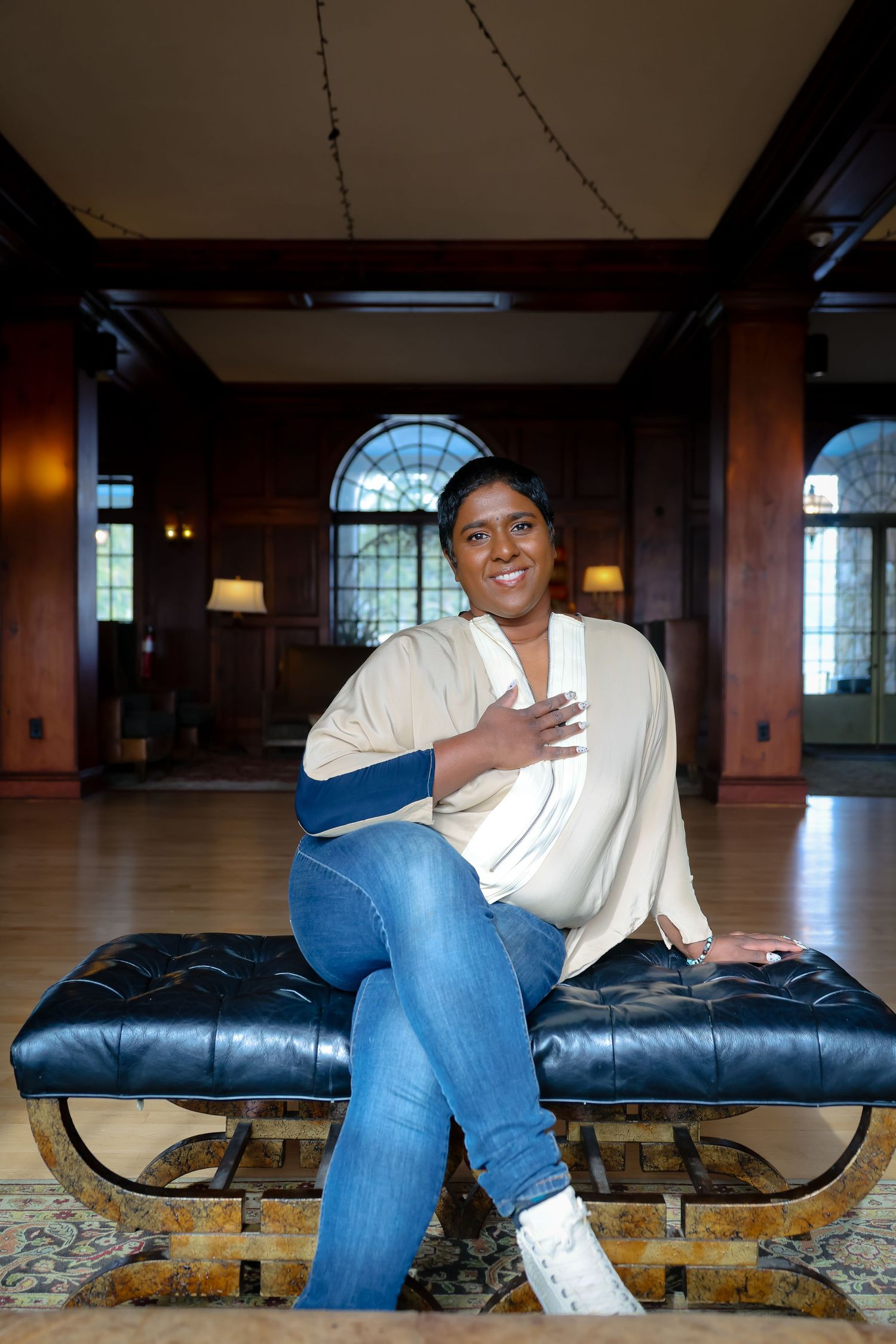 Person wearing jeans and white shoes sits casually on a dark leather bench in a room with wooden details.