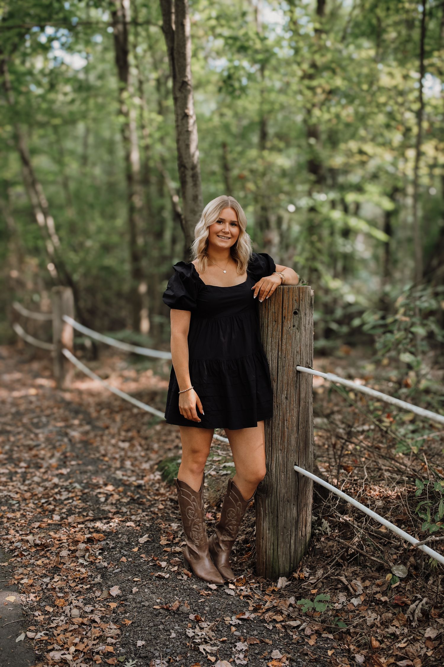 A person in a black dress and brown boots poses casually against a wooden post in a wooded forest setting.