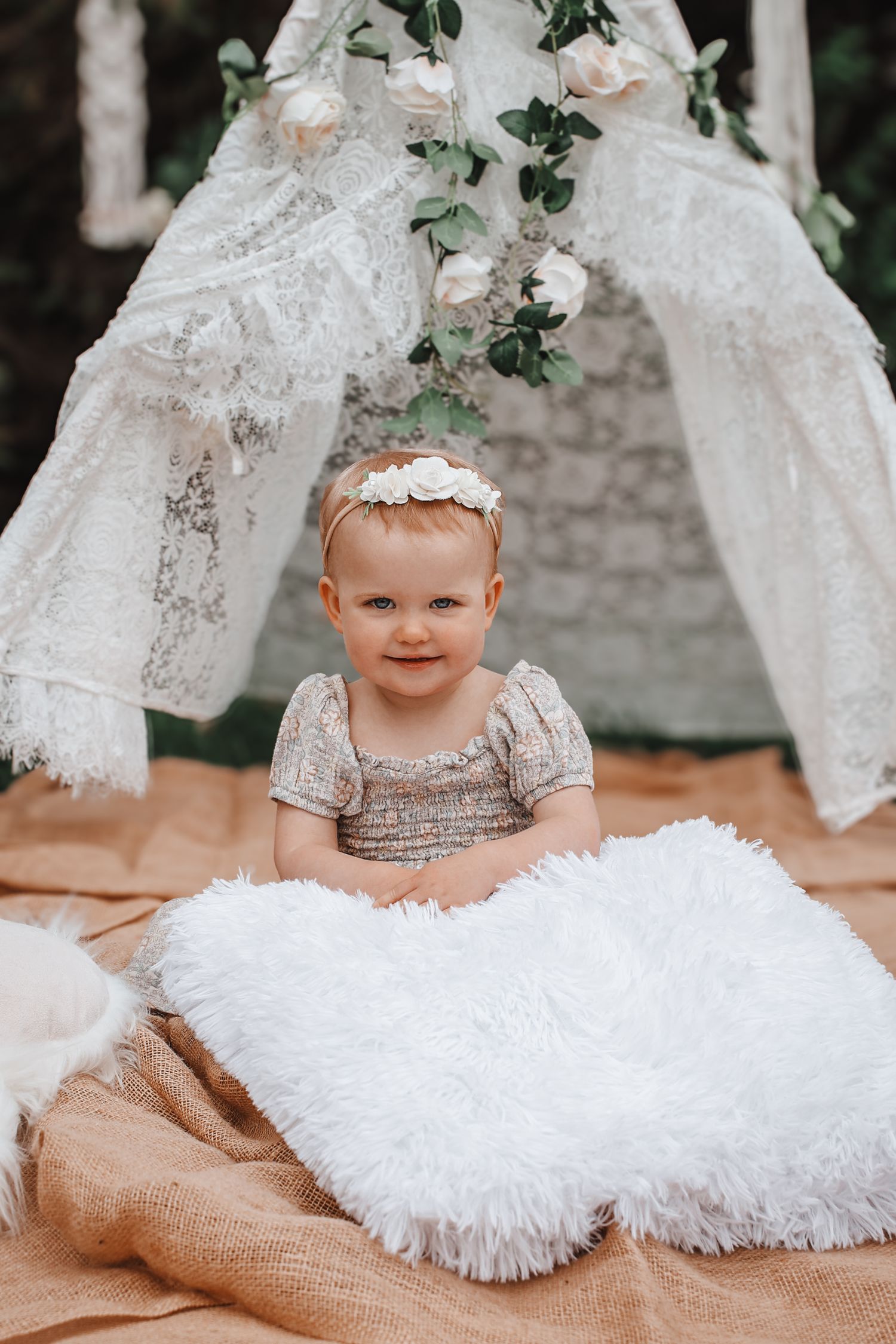 A baby in a silver lace dress and headband poses on a burlap blanket with white tulle decorations and greenery backdrop.