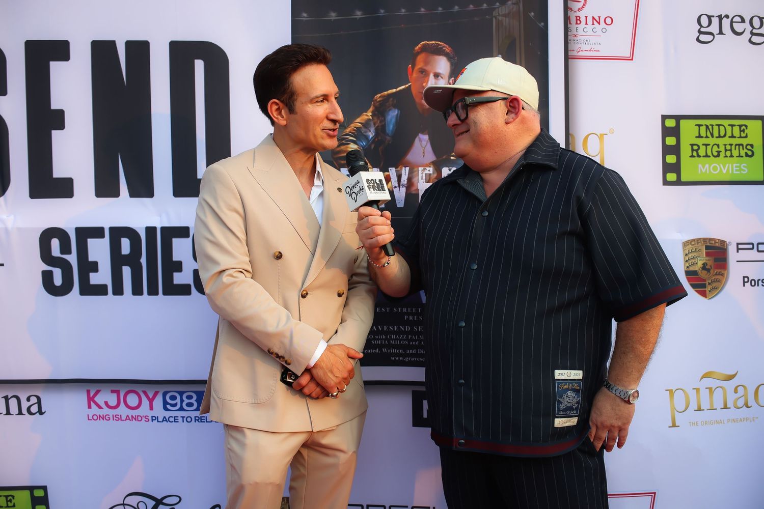 Two men stand together at an event in front of a promotional backdrop with sponsor logos and text.