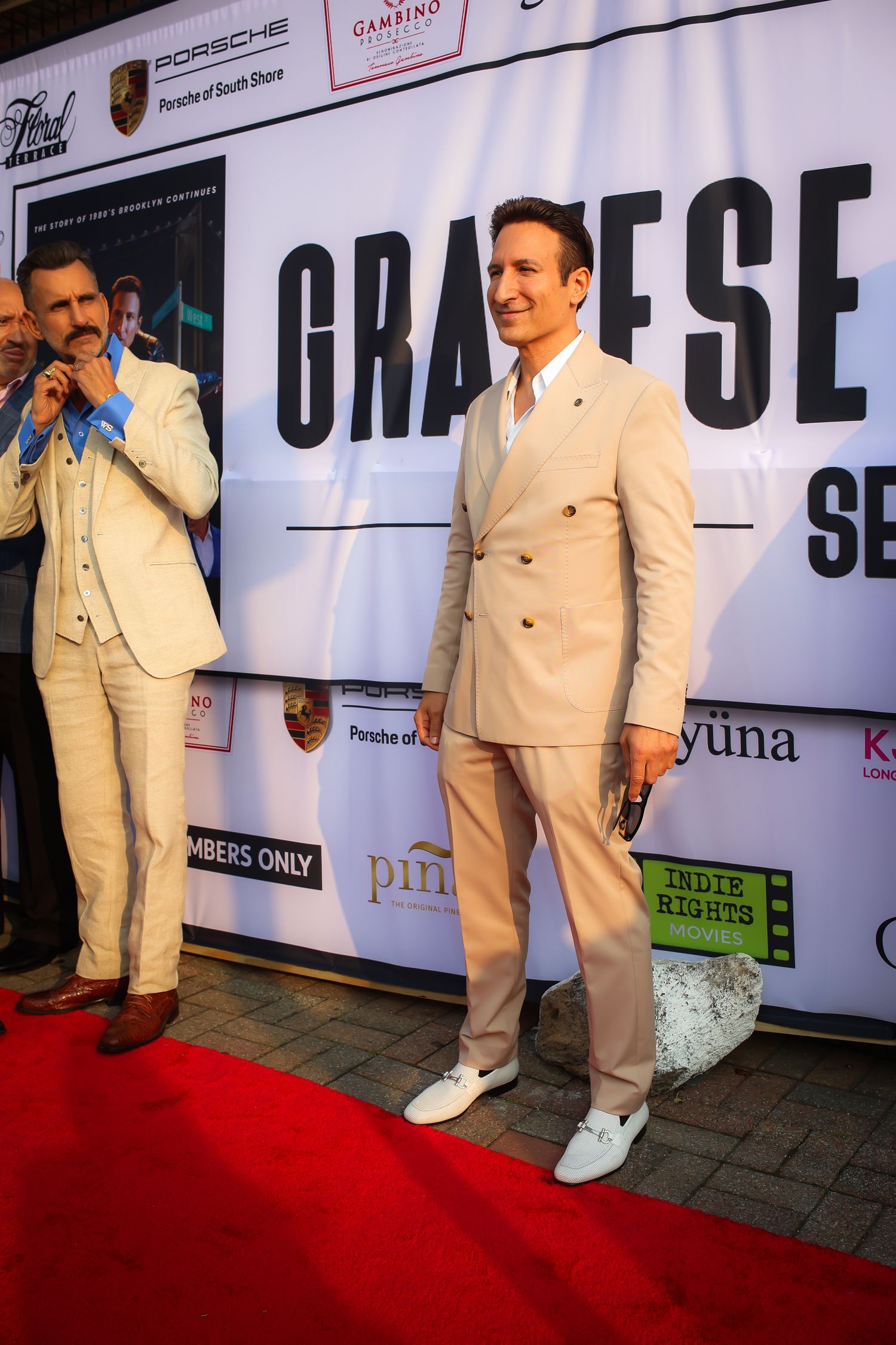 Multiple views of someone posing on a red carpet wearing a cream-colored suit and white shoes at a formal event.