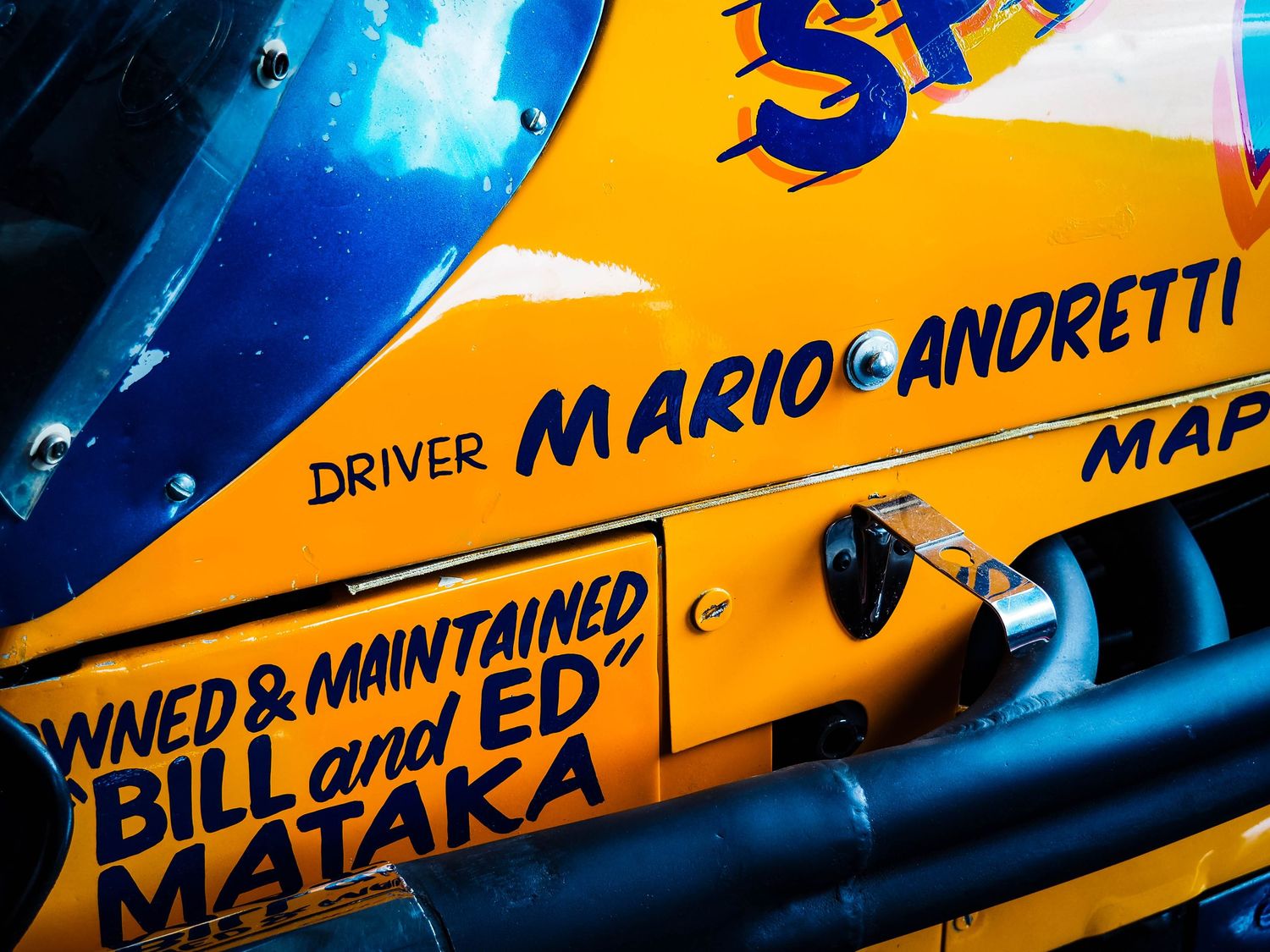 Close-up of a yellow race car with text showing Mario Andretti as driver and Bill and Ed Mataka as owners.
