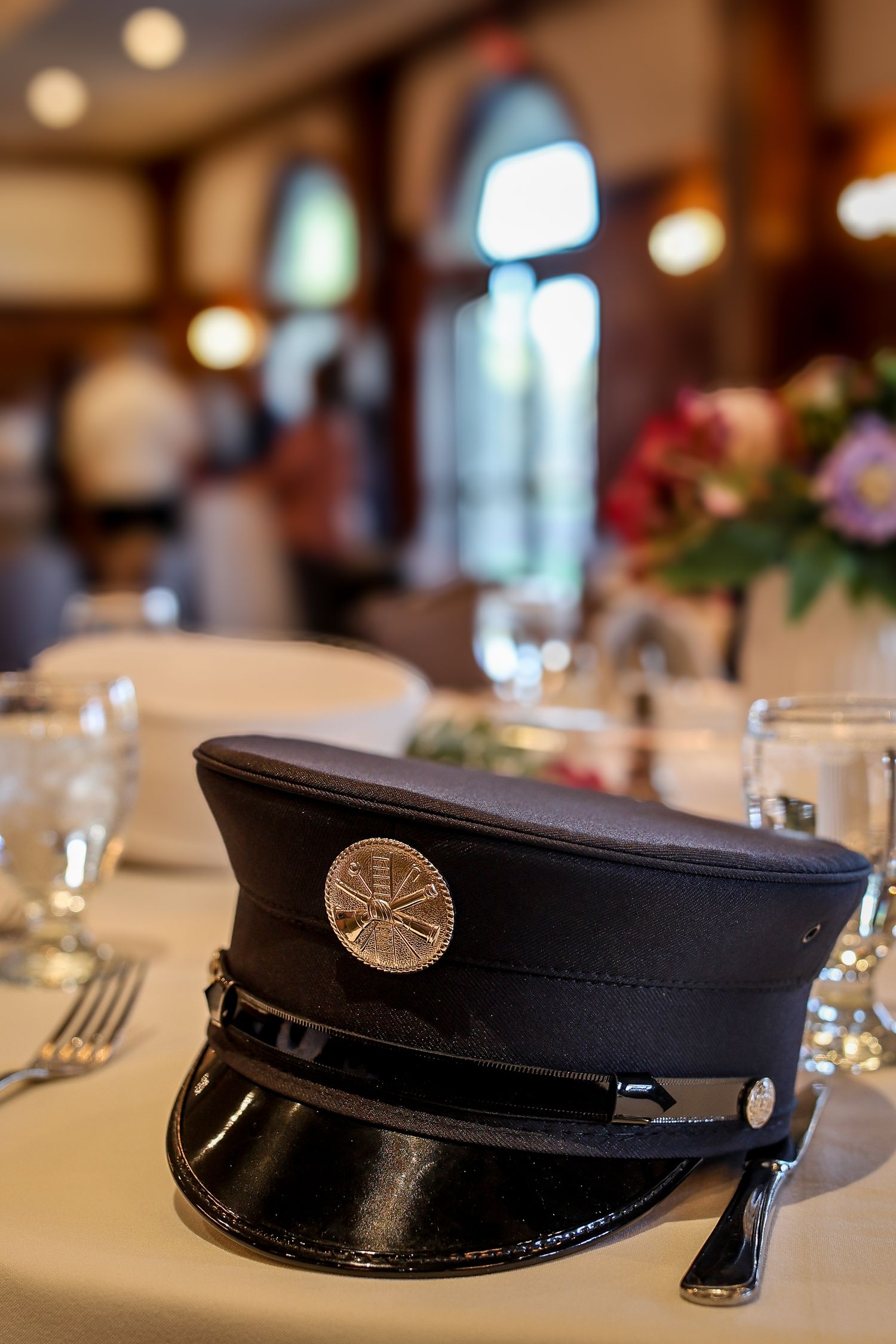 A black police or firefighter's uniform cap sits on a white tablecloth at a formal dining table setting.