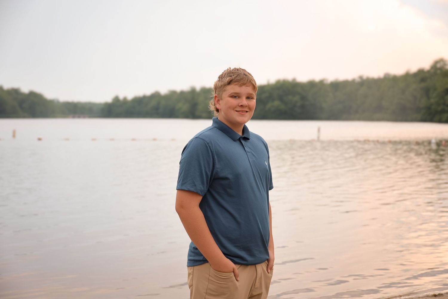 A person in a blue polo shirt stands on a beach at sunset with water and trees in the background.