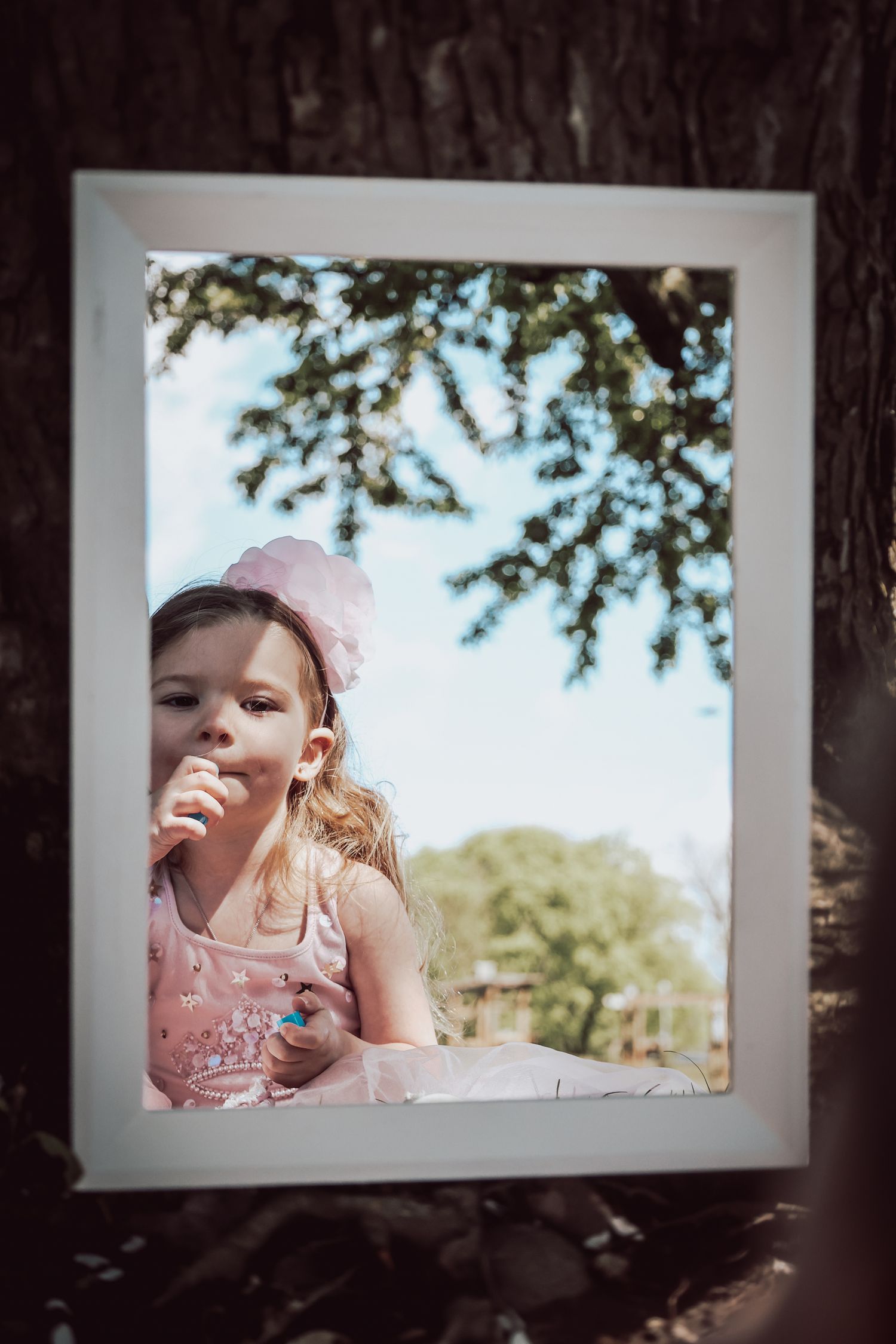 A framed photograph shows a child in a pink dress enjoying a treat outdoors under tree branches on a sunny day.