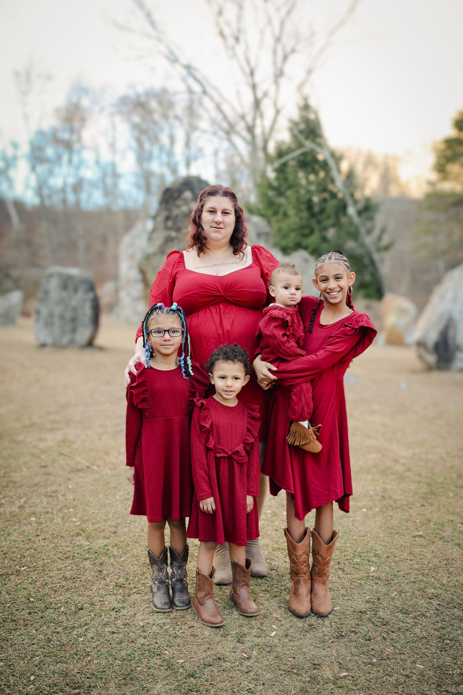 A group in matching burgundy dresses and brown boots pose together outdoors in a grassy field at sunset.