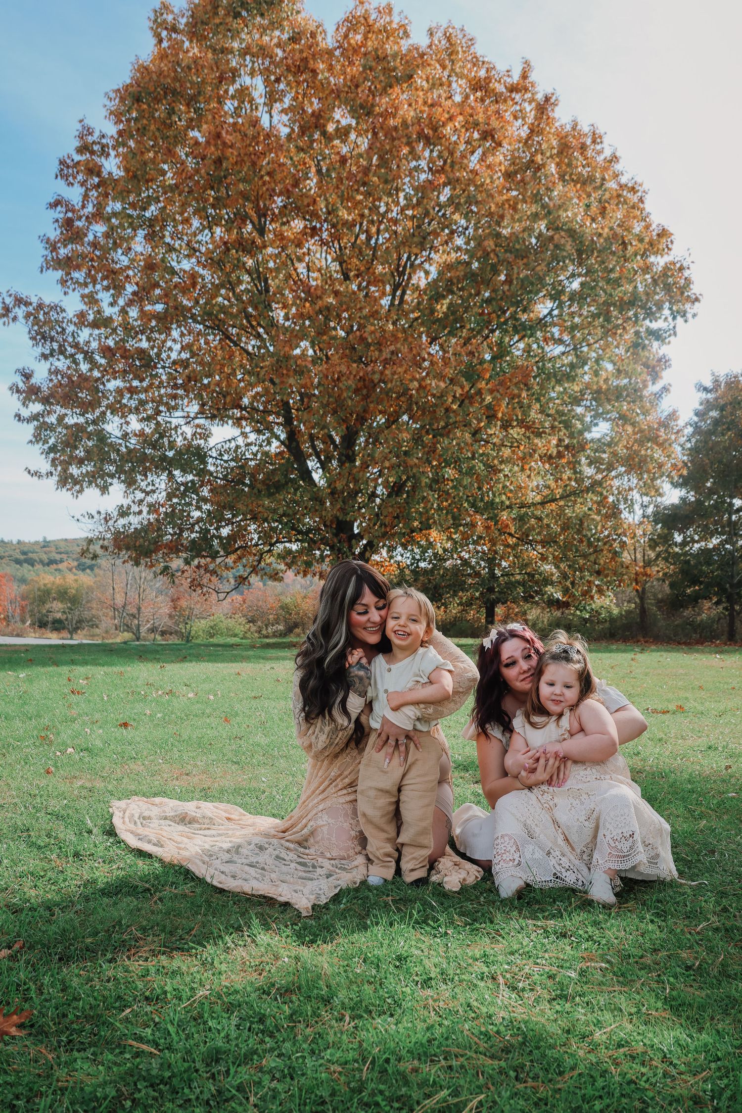 Family relaxing together on blanket under autumn tree in grassy field during outdoor photo session.