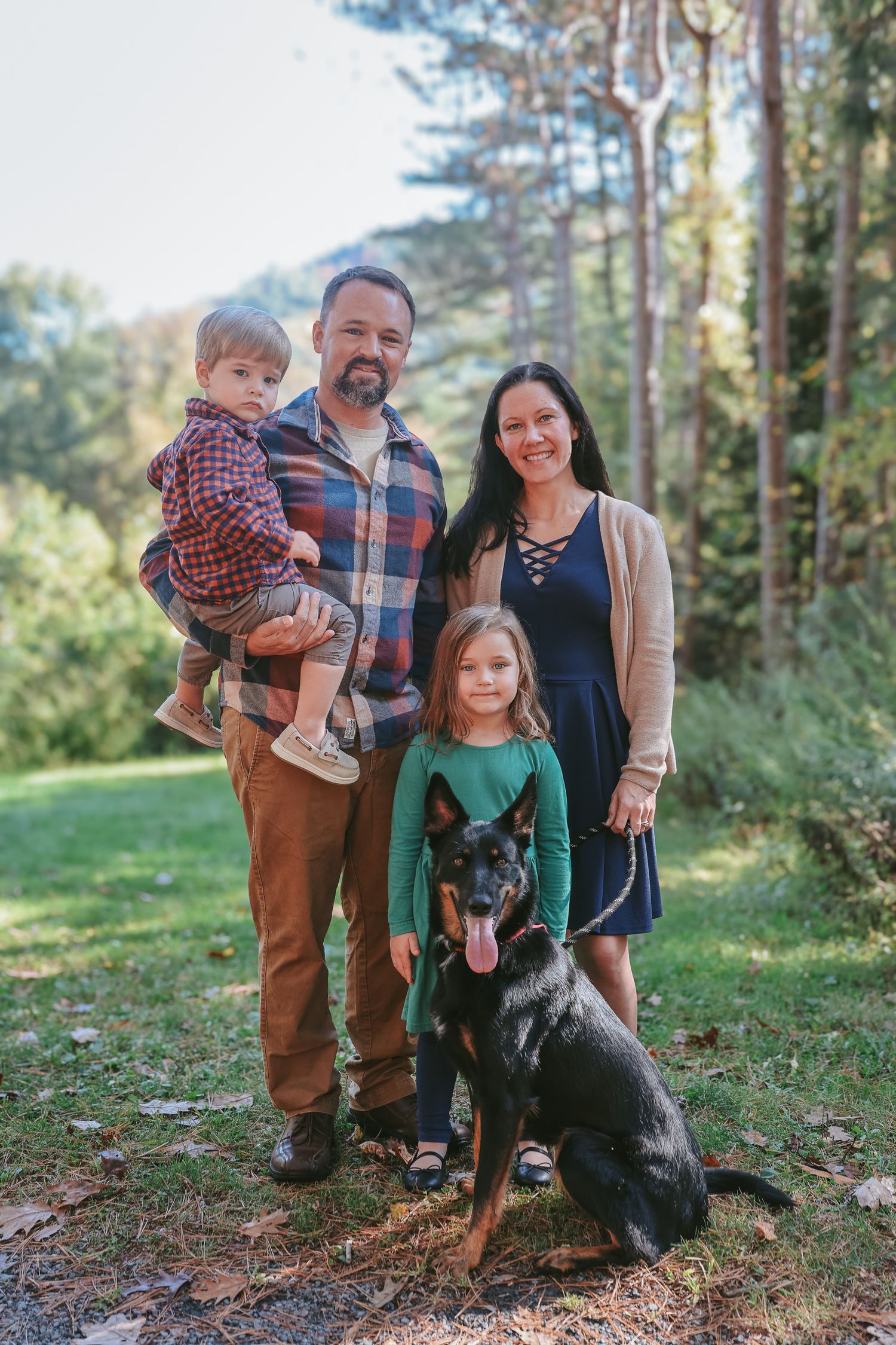 A family poses outdoors in a wooded setting with their black dog on a sunny autumn day.