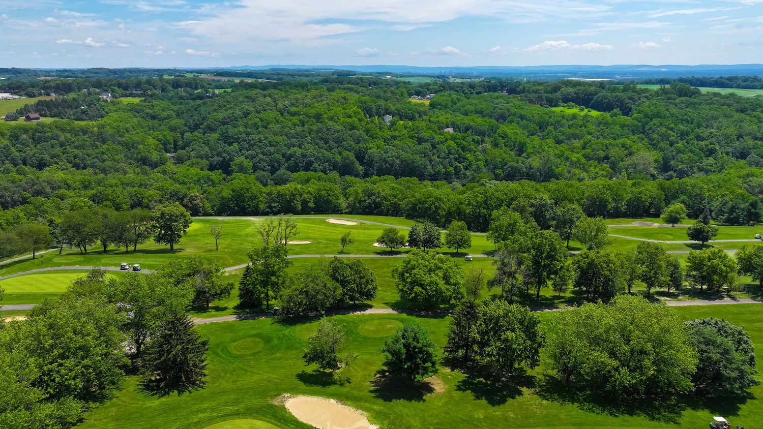 Aerial view of a lush golf course with rolling fairways surrounded by dense forest and trees under blue skies.