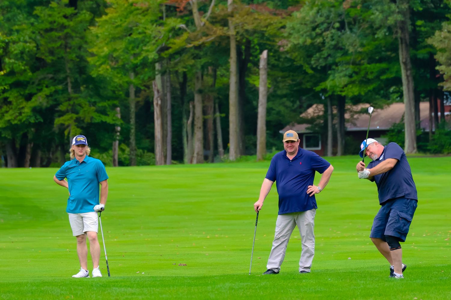 Three golfers stand on a lush green fairway surrounded by tall trees during a round of golf.