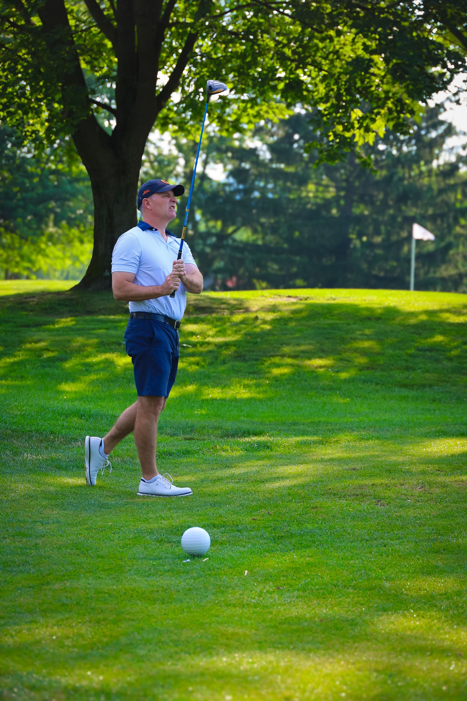 A golfer in white polo and navy shorts stands on a lush green course with a golf ball and flag in the distance.