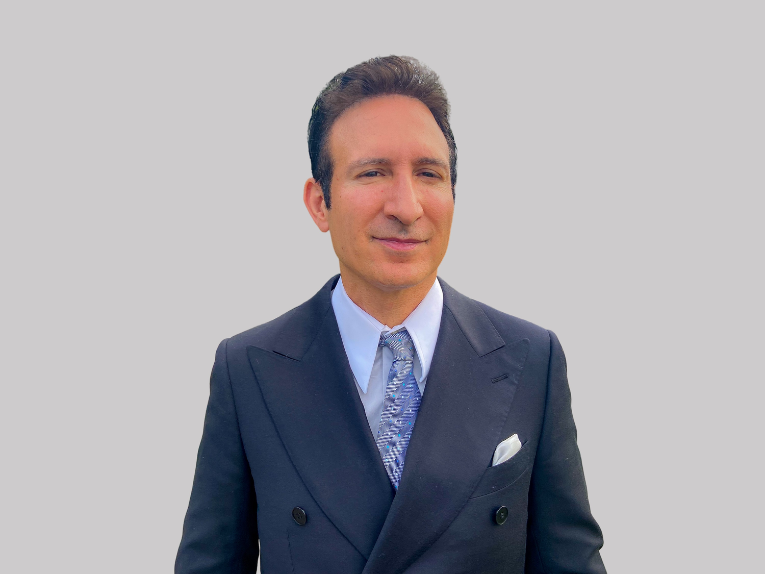 A person in a navy suit and light blue tie stands against a neutral background in a professional business portrait.