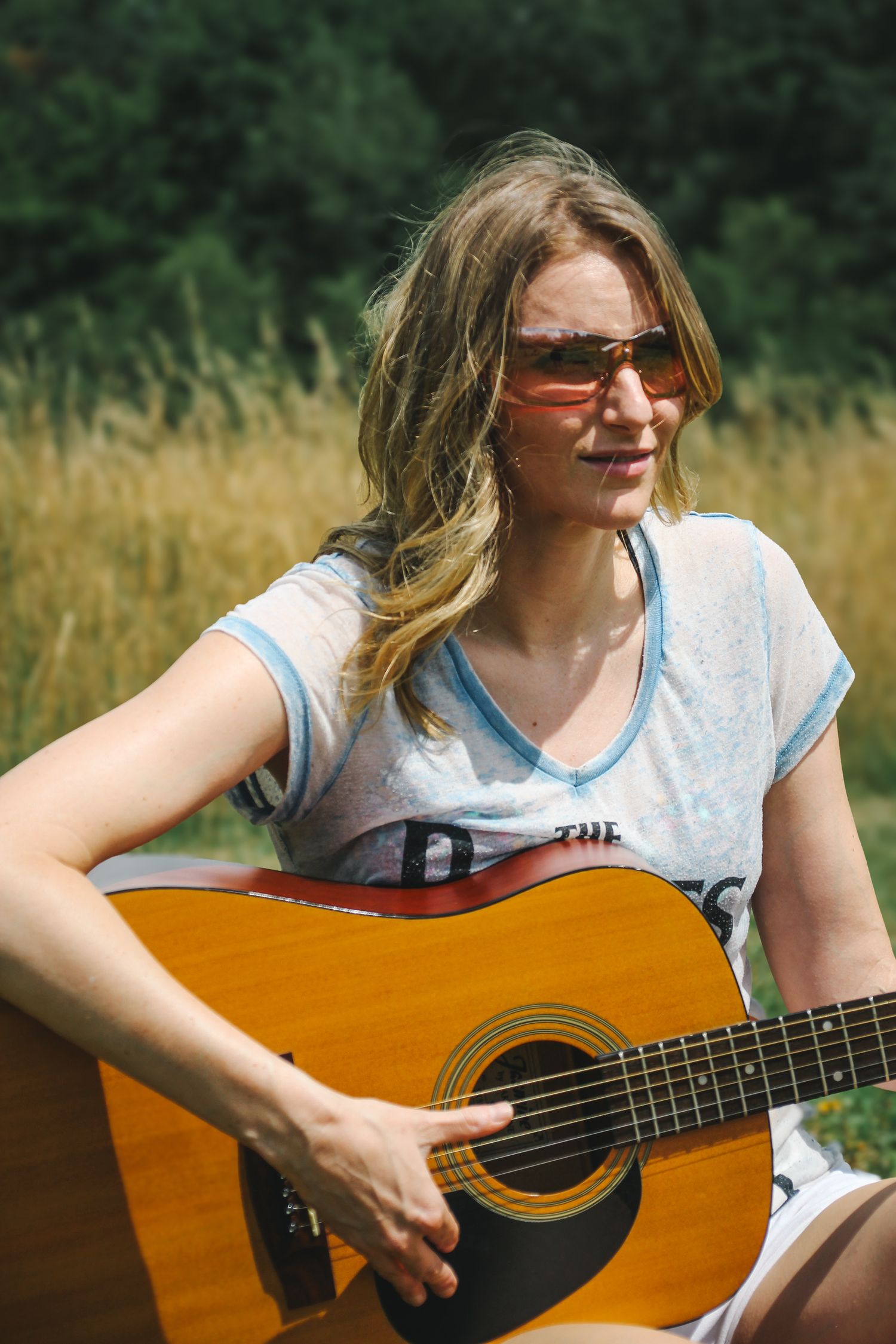 A person in a white t-shirt plays acoustic guitar outdoors in a sunny field with tall grass in the background.