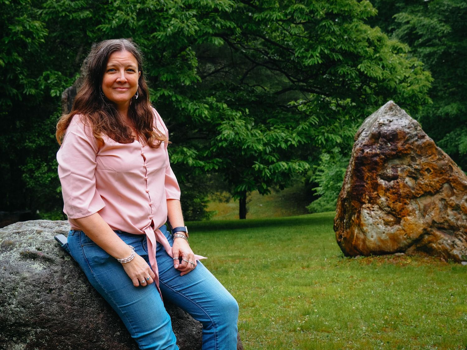 A person in a pink top and jeans sits on a large rock formation in a lush green park setting.