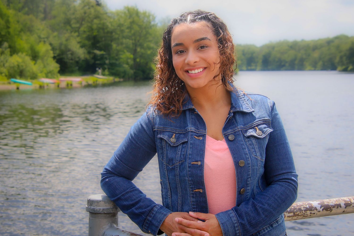 Person in denim jacket smiling near a scenic lake on a sunny day with trees in the background.