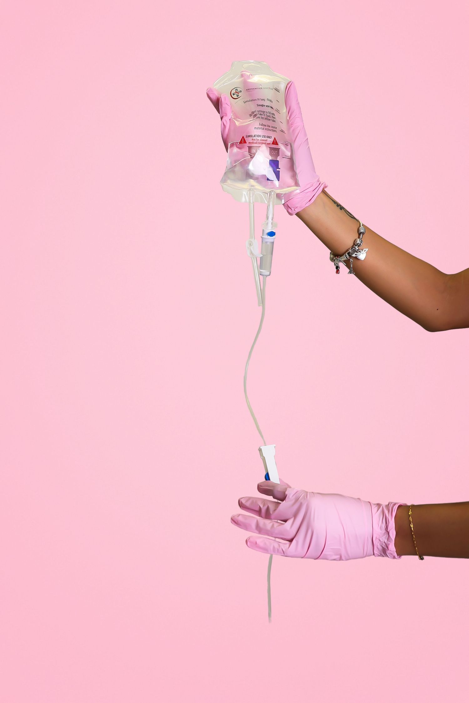 Hands in pink gloves holding an IV drip bag and tubing against a pink background.