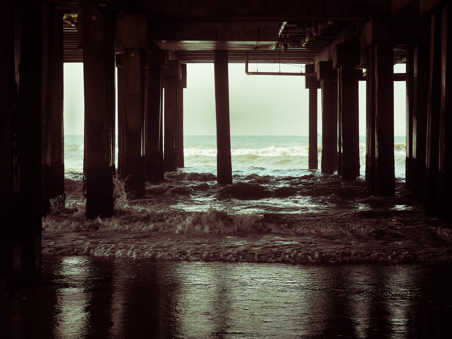 Moody ocean waves crashing under a dark wooden pier with dramatic stormy skies visible in the background.