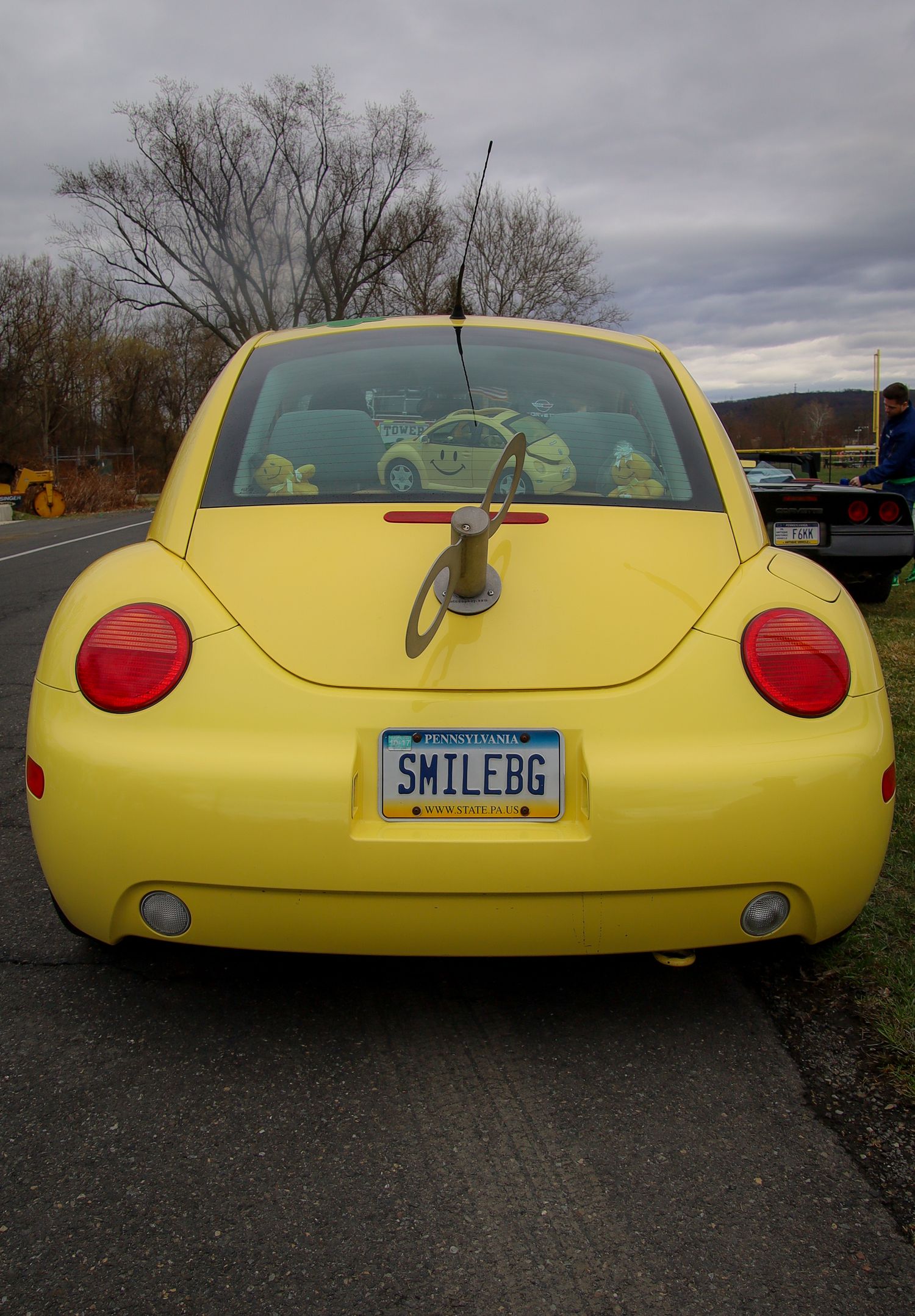 A bright yellow Volkswagen Beetle with red taillights and a custom license plate reading SMILEBG parked on a cloudy day.