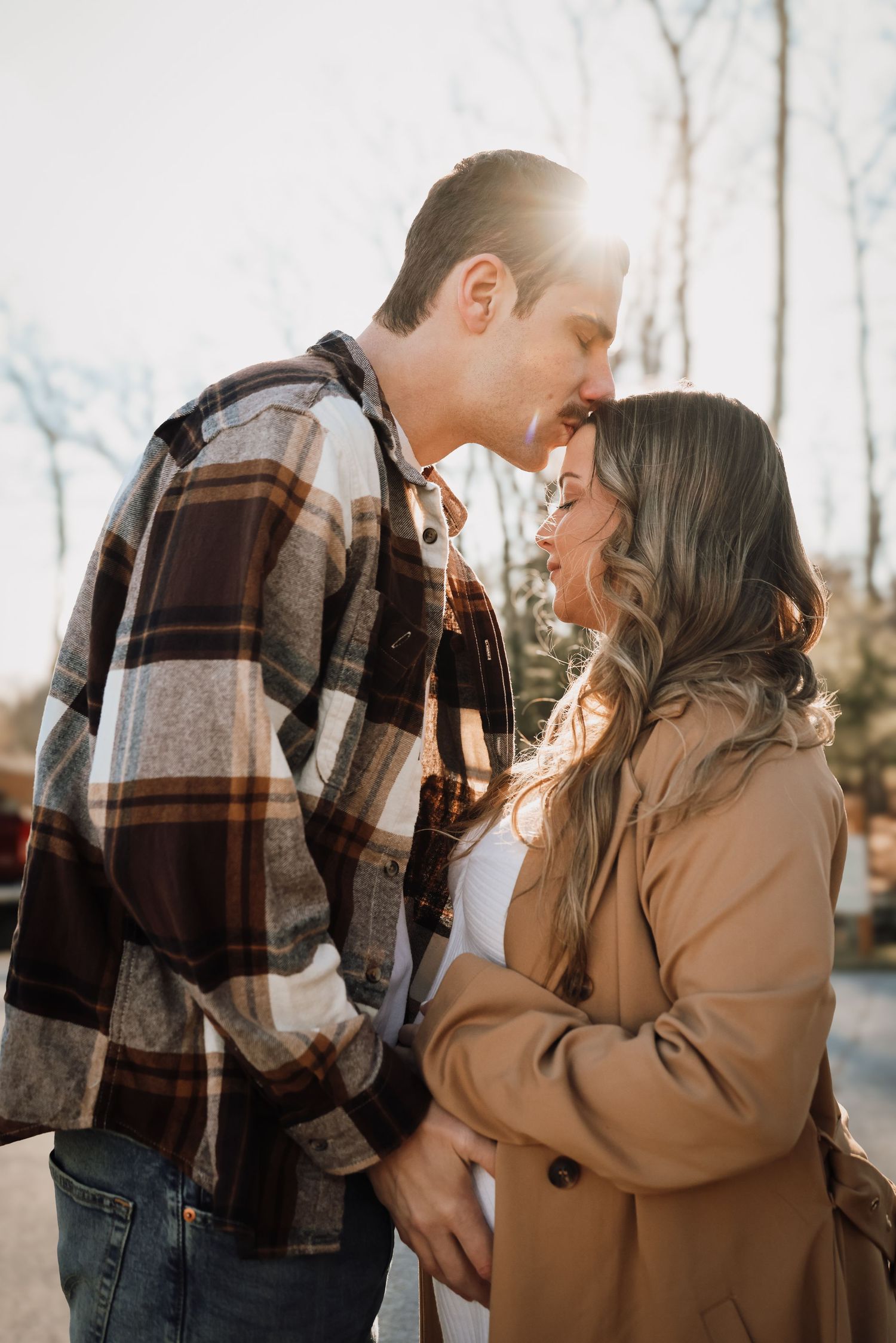 A romantic winter engagement photo with a couple in plaid and tan coats sharing an intimate moment in sunlit woods.