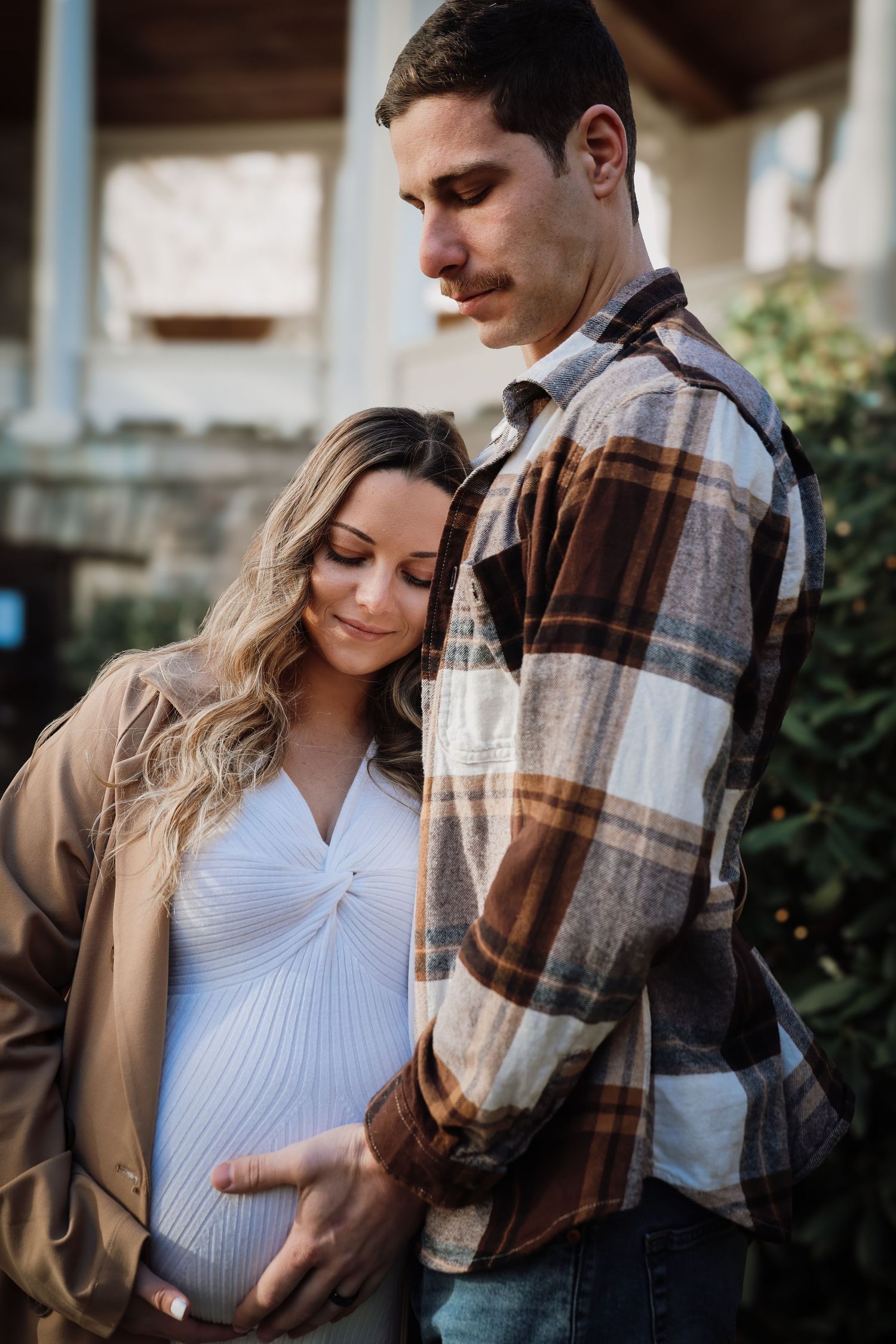 A couple shares an intimate moment outdoors as they await their new arrival, with her wearing white and him in plaid.