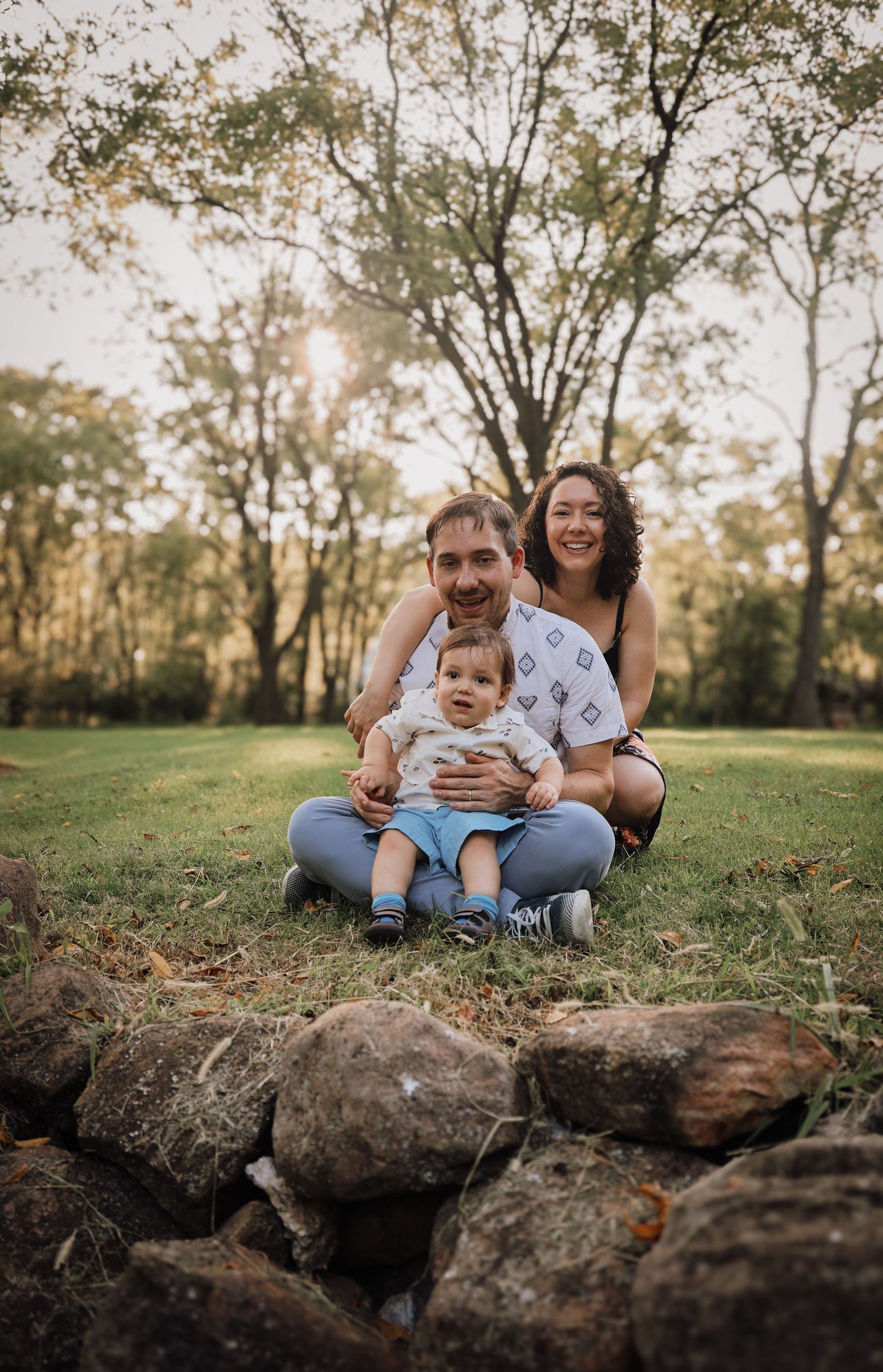 Family sitting together on grassy hill during golden hour sunset with rocks in foreground.