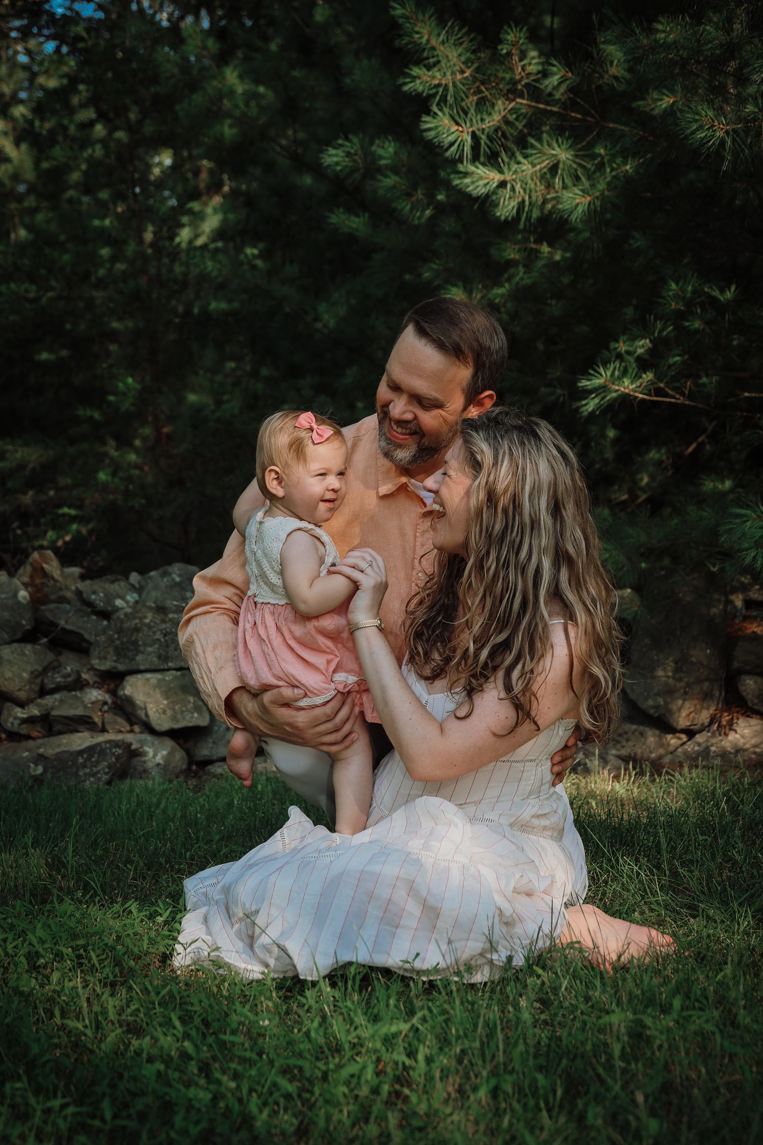 A family shares a joyful moment together while sitting on grass near pine trees in warm evening light.