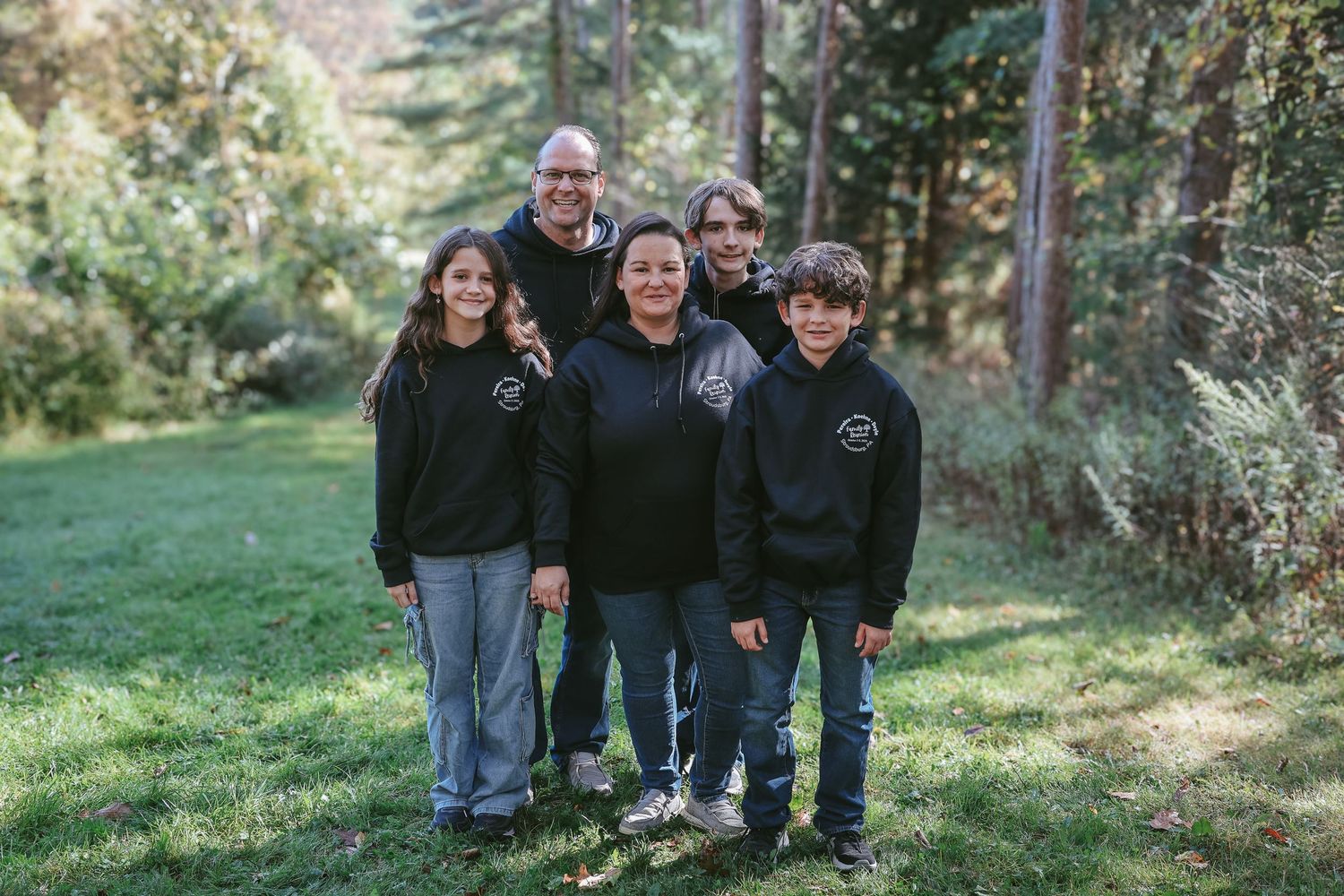 A group in dark clothing poses together outdoors on a grassy hillside surrounded by evergreen trees.
