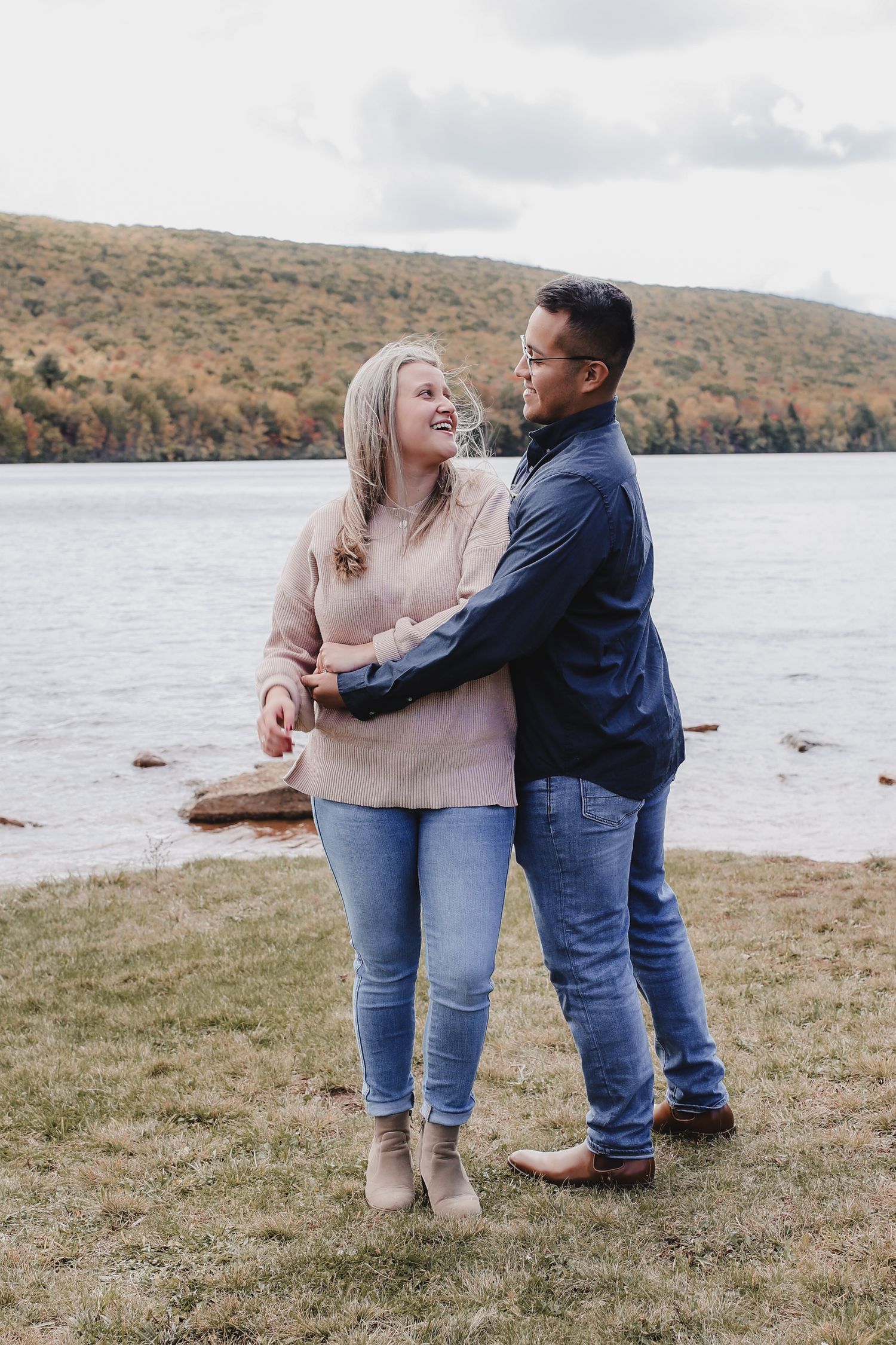 A couple shares a joyful moment together by a lake with autumn hills in the background.