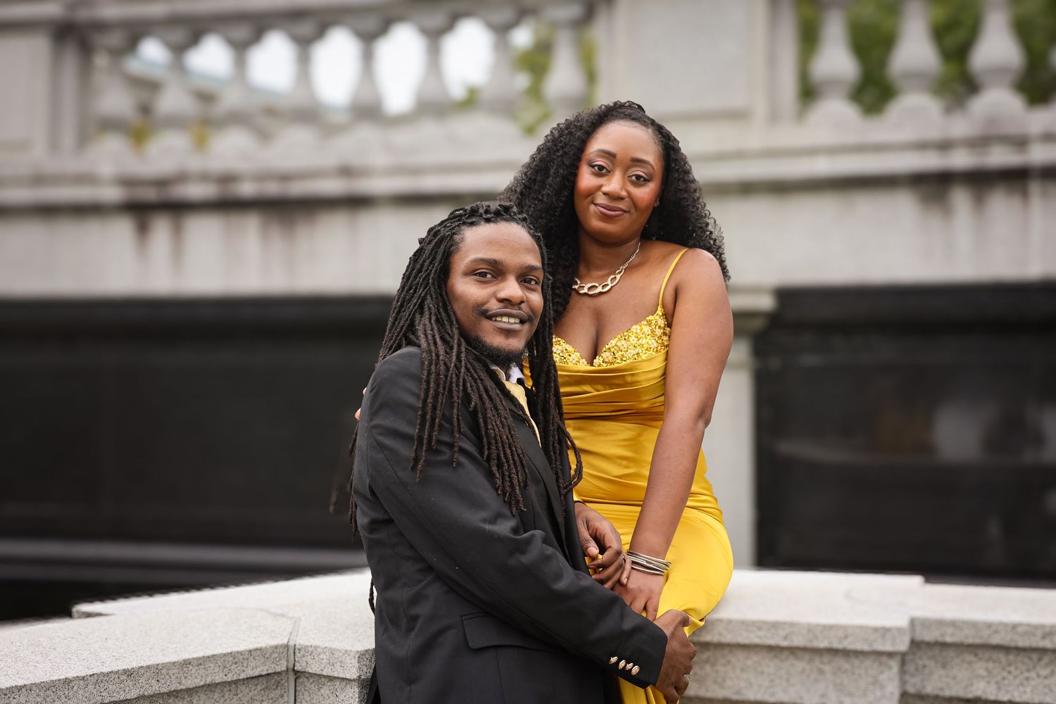 A couple sits together on outdoor stone steps, with one person wearing a yellow dress and the other in dark formal attire.