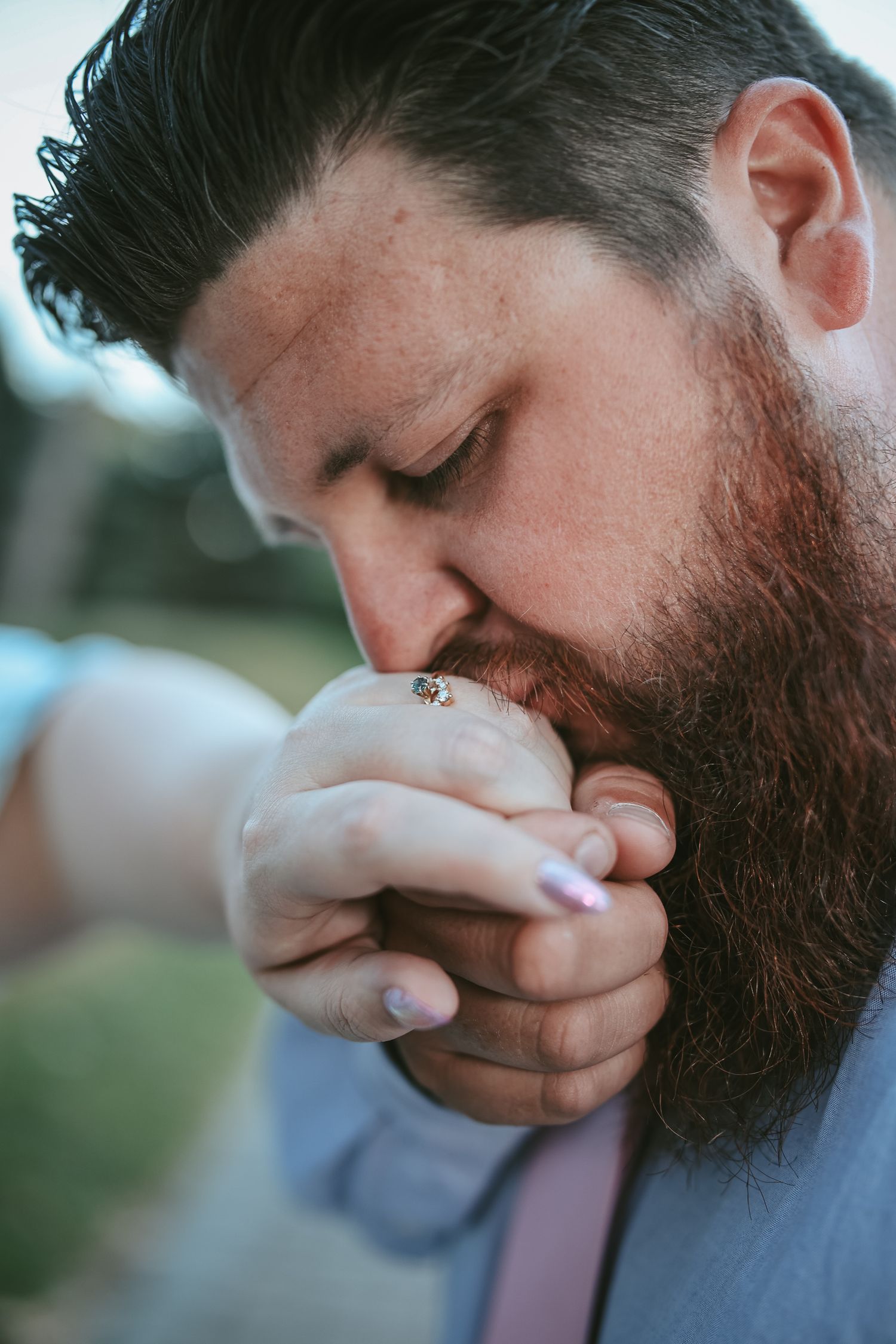 Close-up photo of a bearded person in a suit gently kissing hands with closed eyes in an emotional moment.