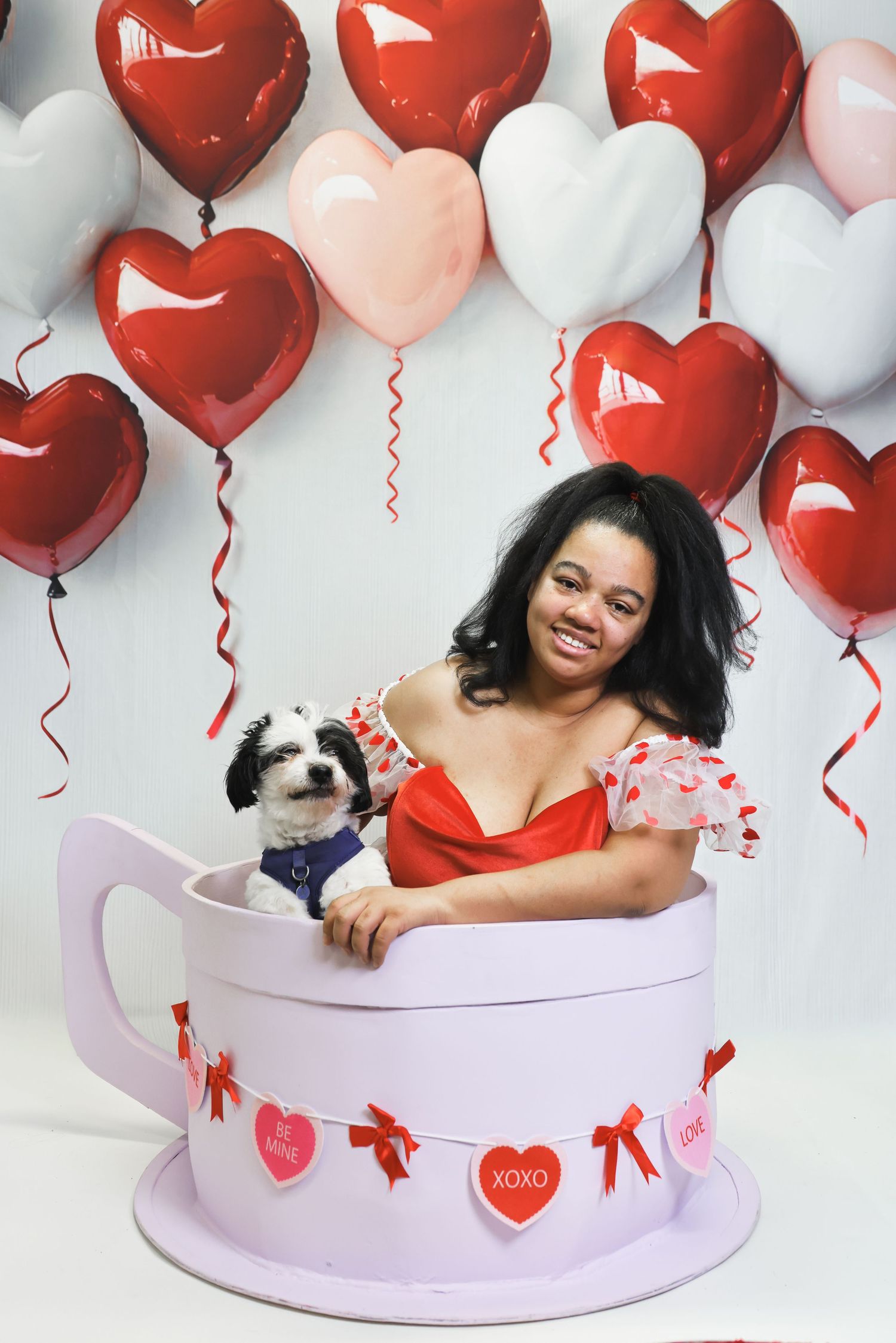 A Valentine's Day photo setup with a person and small dog in a pink round prop surrounded by red heart balloons.