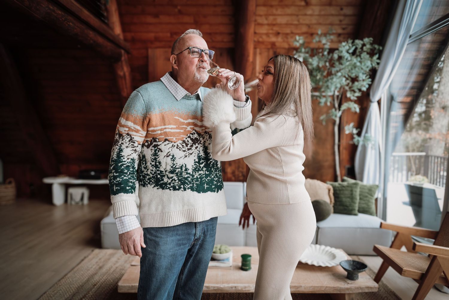 A couple in casual winter outfits embraces in a rustic indoor setting with wooden walls and white floral decor.